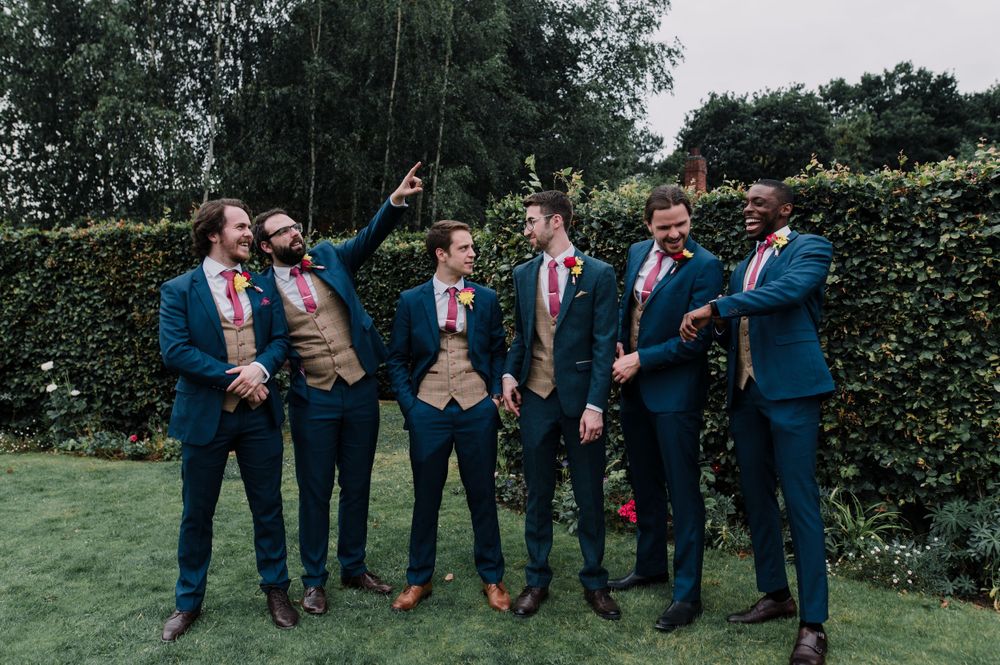 Wedding party in blue suits posing against garden hedge backdrop.