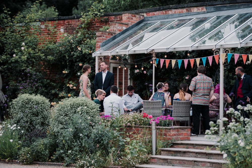 Guests gather under a decorative canopy with colorful bunting in a garden setting during a celebration.