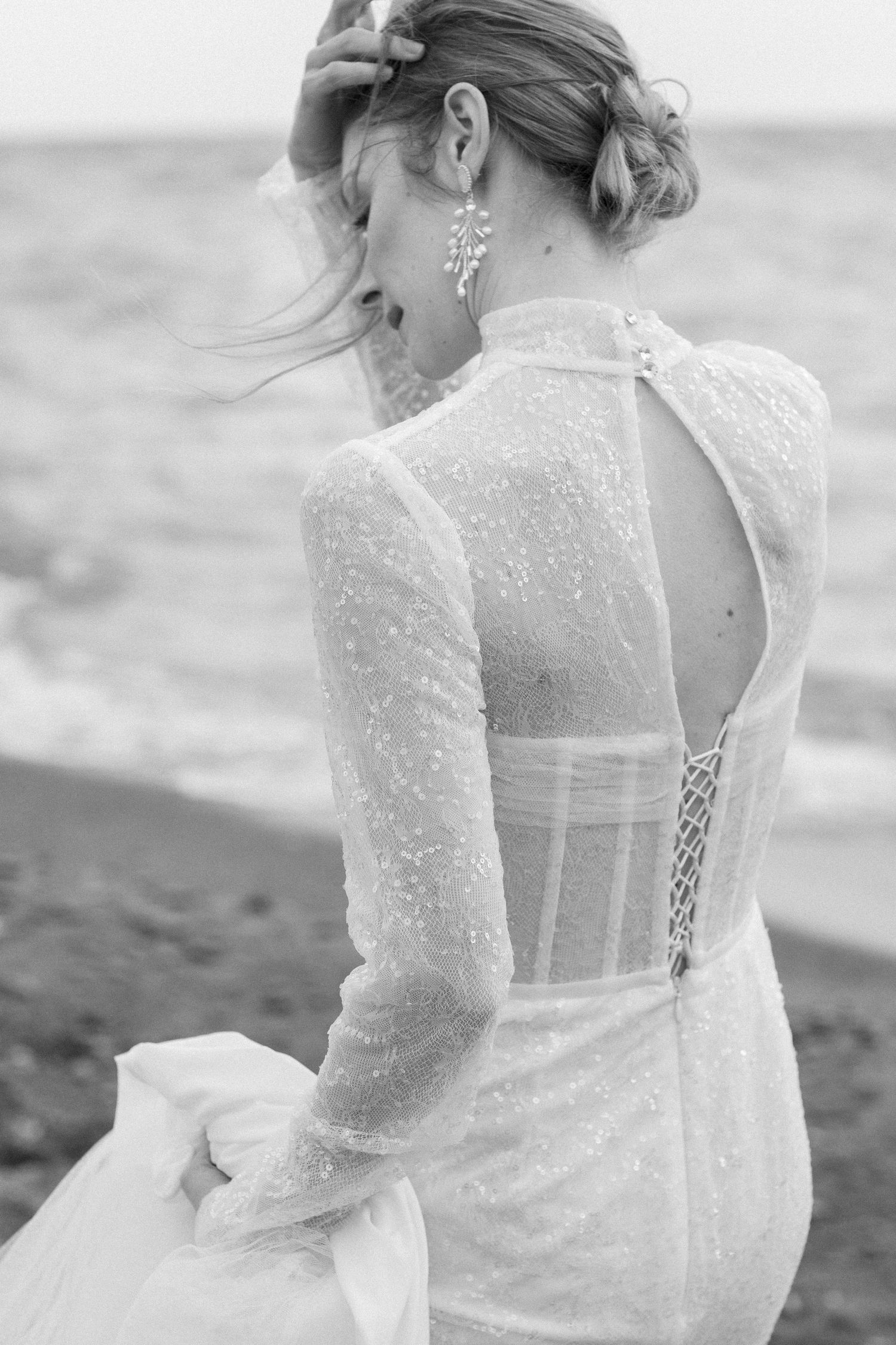 Ethereal lace wedding dress with sheer long sleeves and open back detail shown against a beach backdrop.