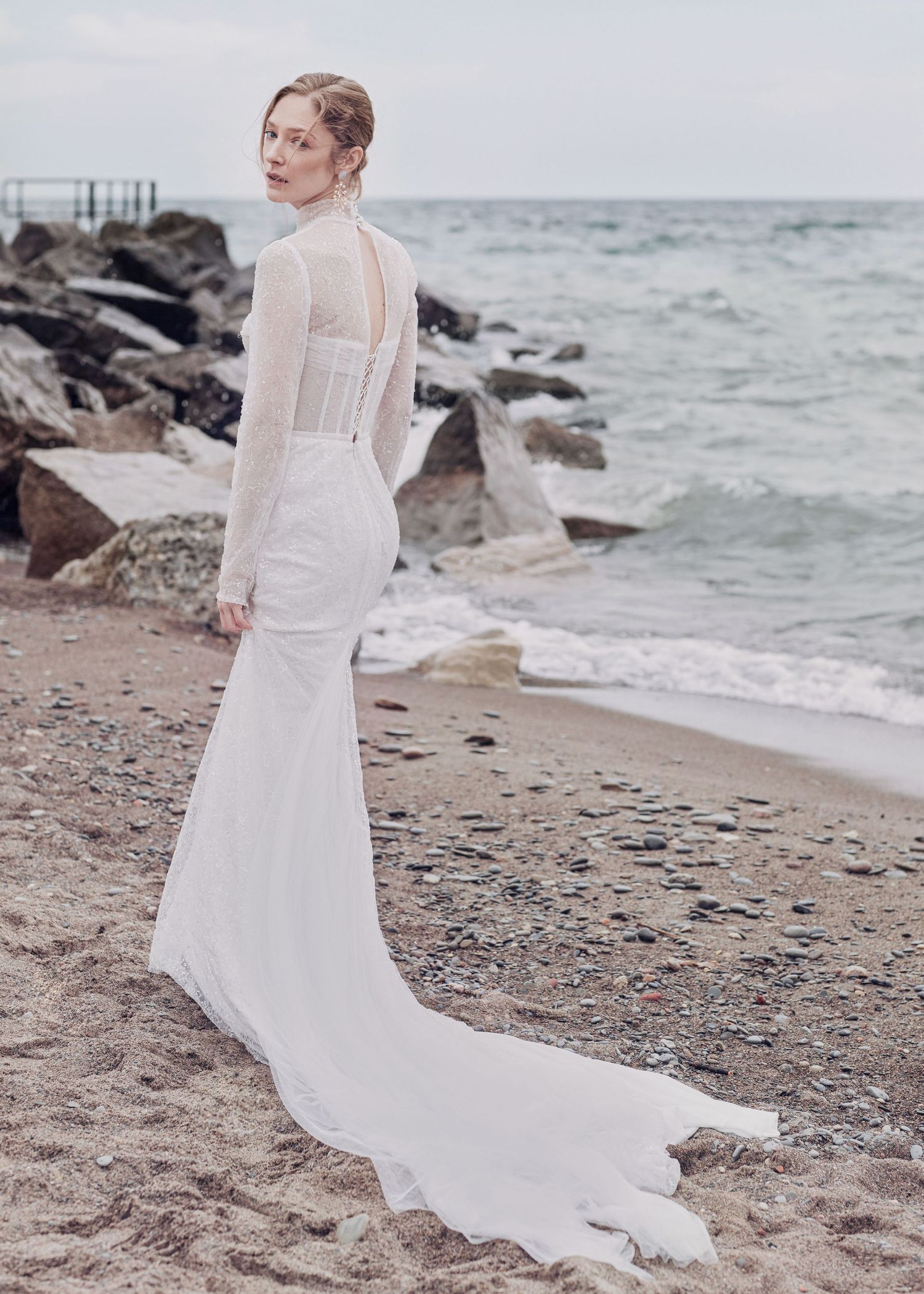 Elegant fitted wedding gown with sheer details and dramatic train photographed on rocky beach.