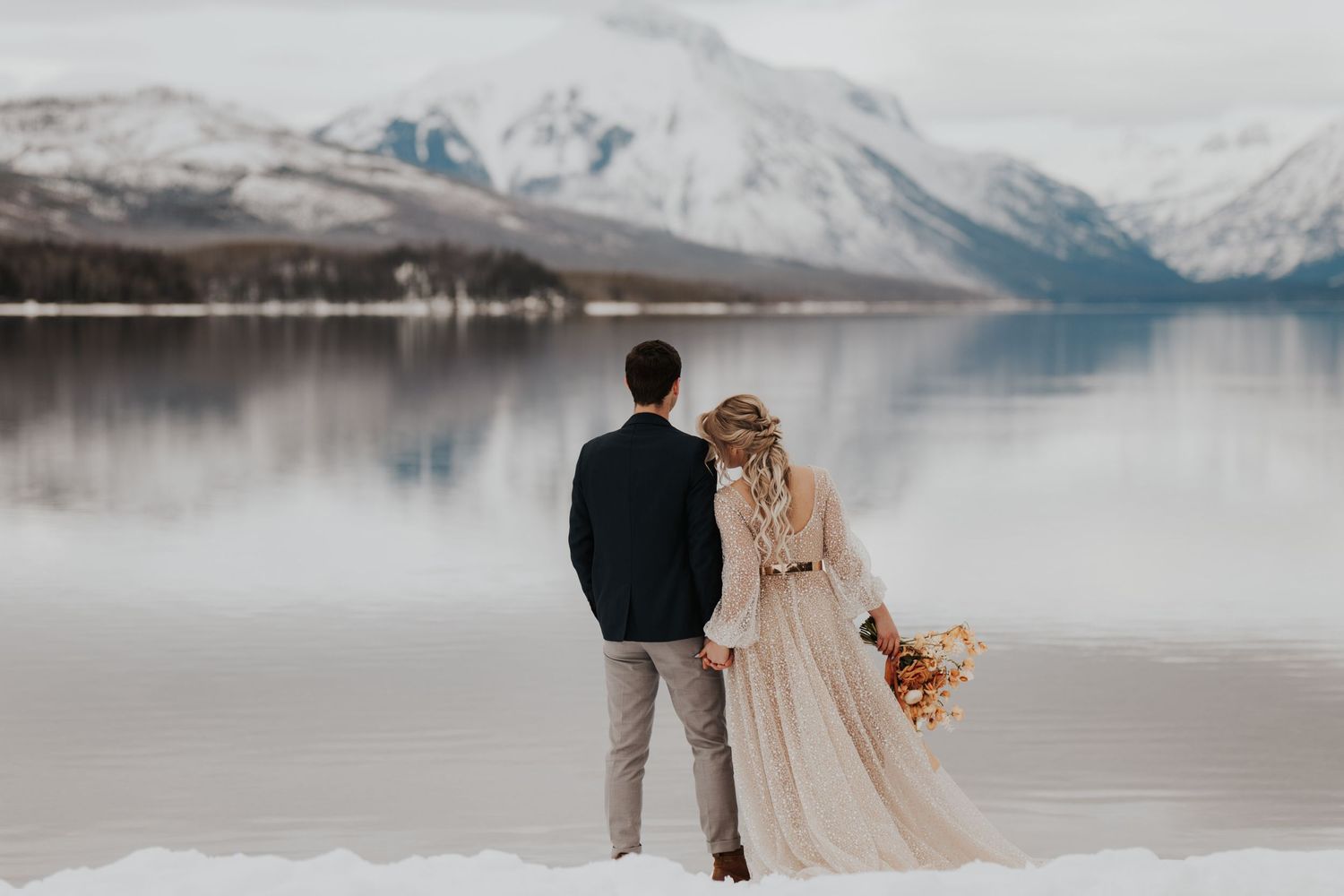 Bride and groom who are eloping at GNP overlook the smooth lake and snow covered mountains in their weddign attire