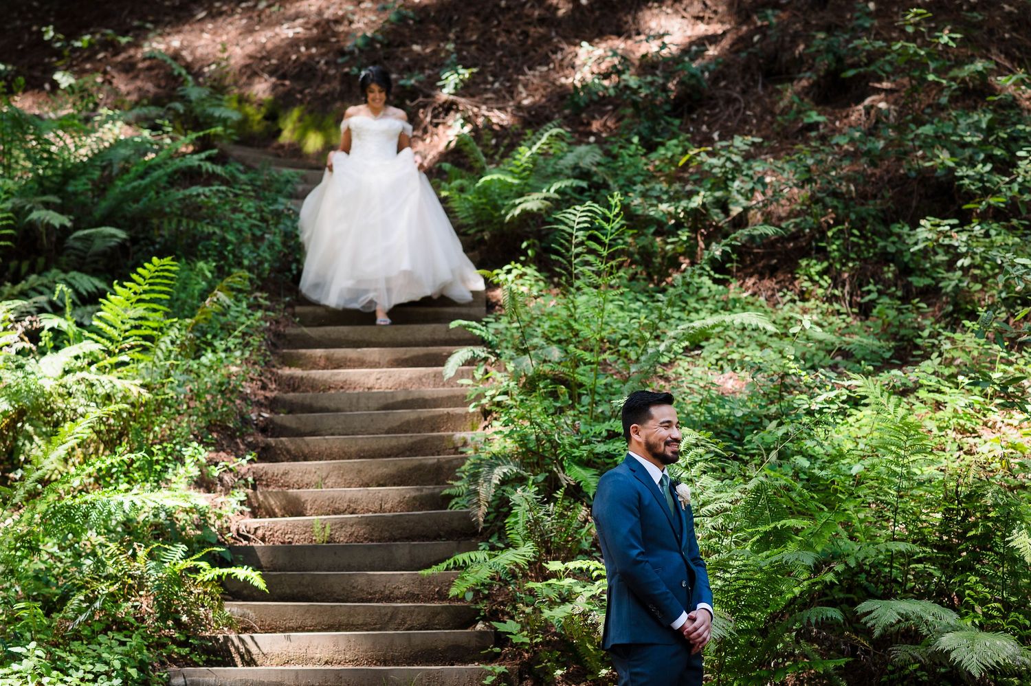 A romantic first look moment on rustic forest stairs surrounded by green ferns.