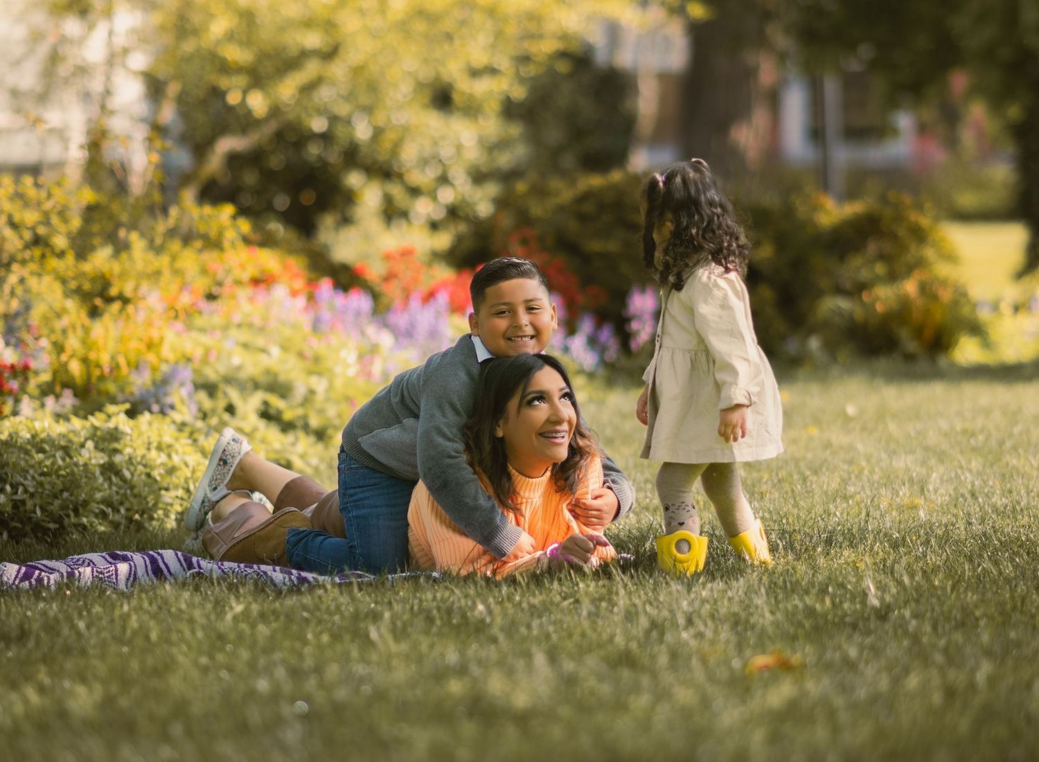 Mom laying down with son on top of her posing. Littler girl with yellow rain boots looking down at mom being playful.