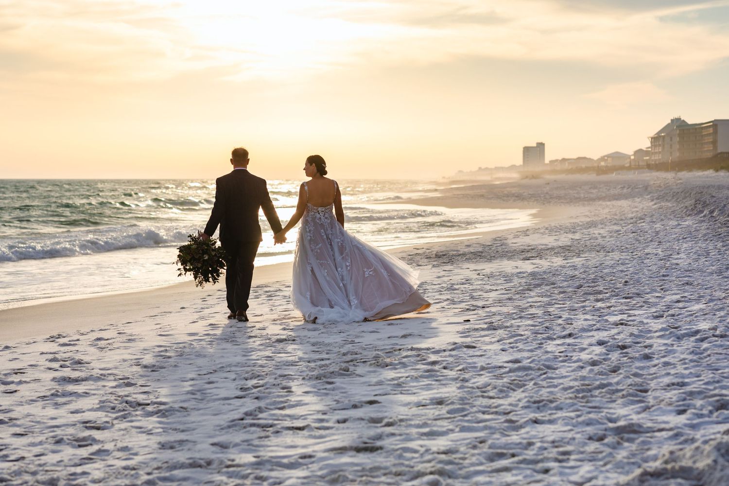 Silhouetted couple walking hand in hand on beach at sunset in wedding attire.
