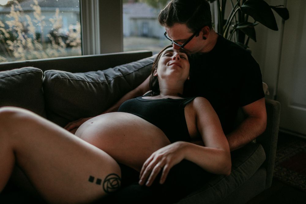 pregnant person lays in partners lap on couch and recieves forehead kiss portland oregon maternity photographer