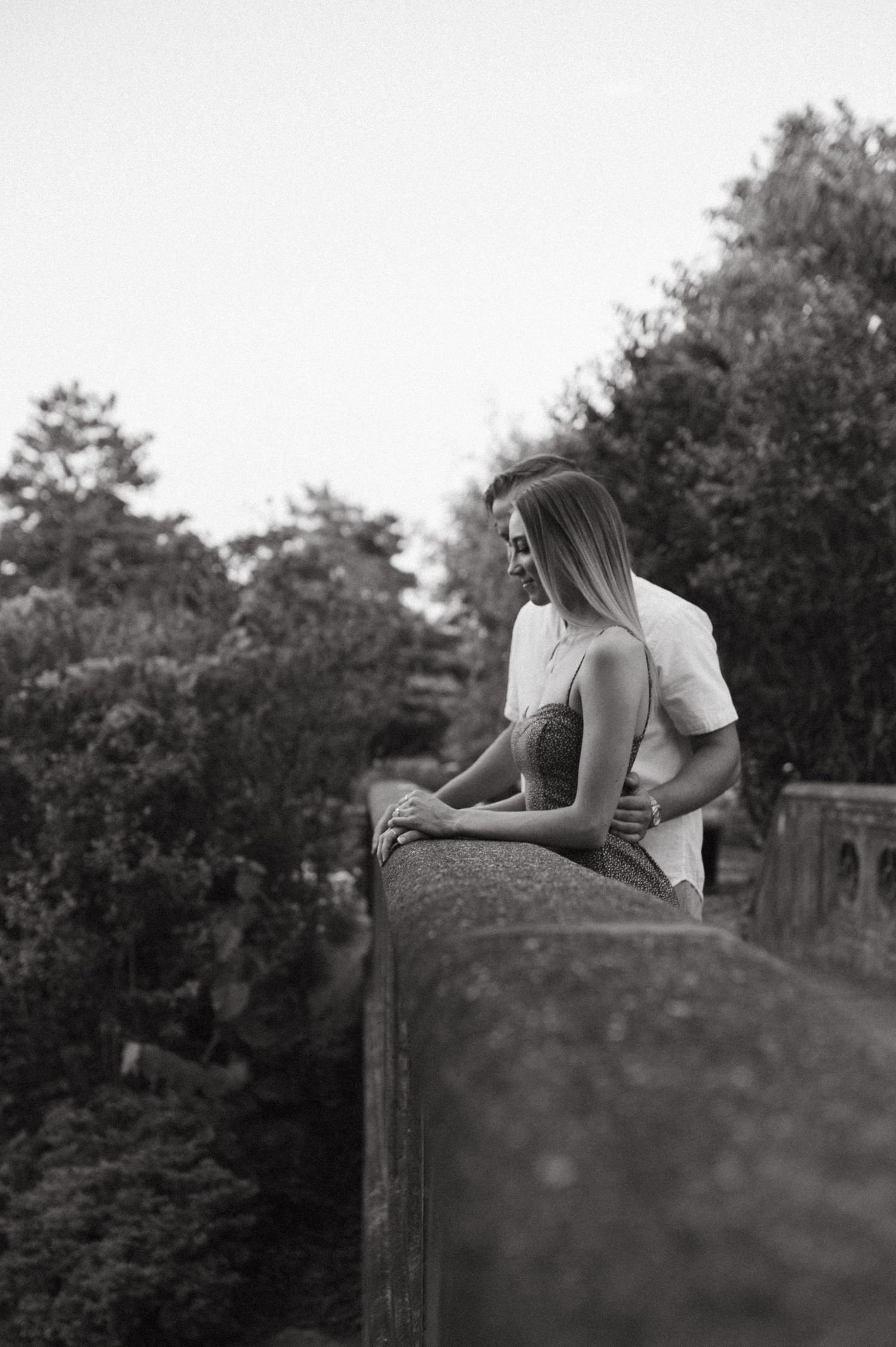 Black and white photo of couple sharing intimate moment on stone bridge overlook.