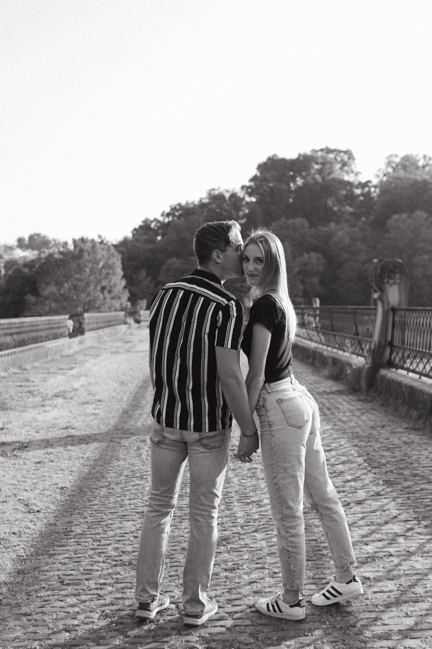 Black and white photo of a couple in casual wear standing on a historic bridge pathway.