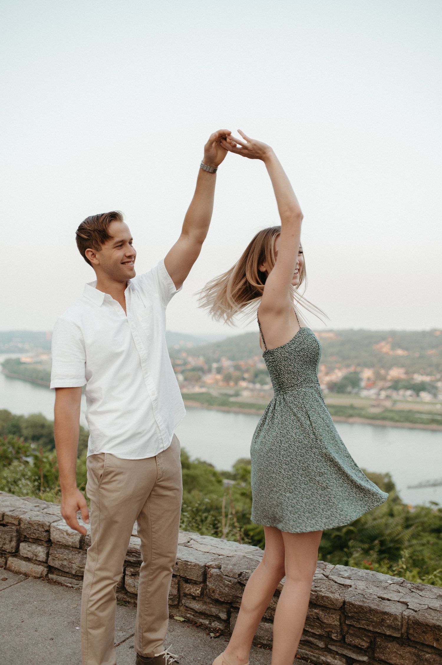 Couple dancing and twirling on stone wall overlooking scenic river valley.