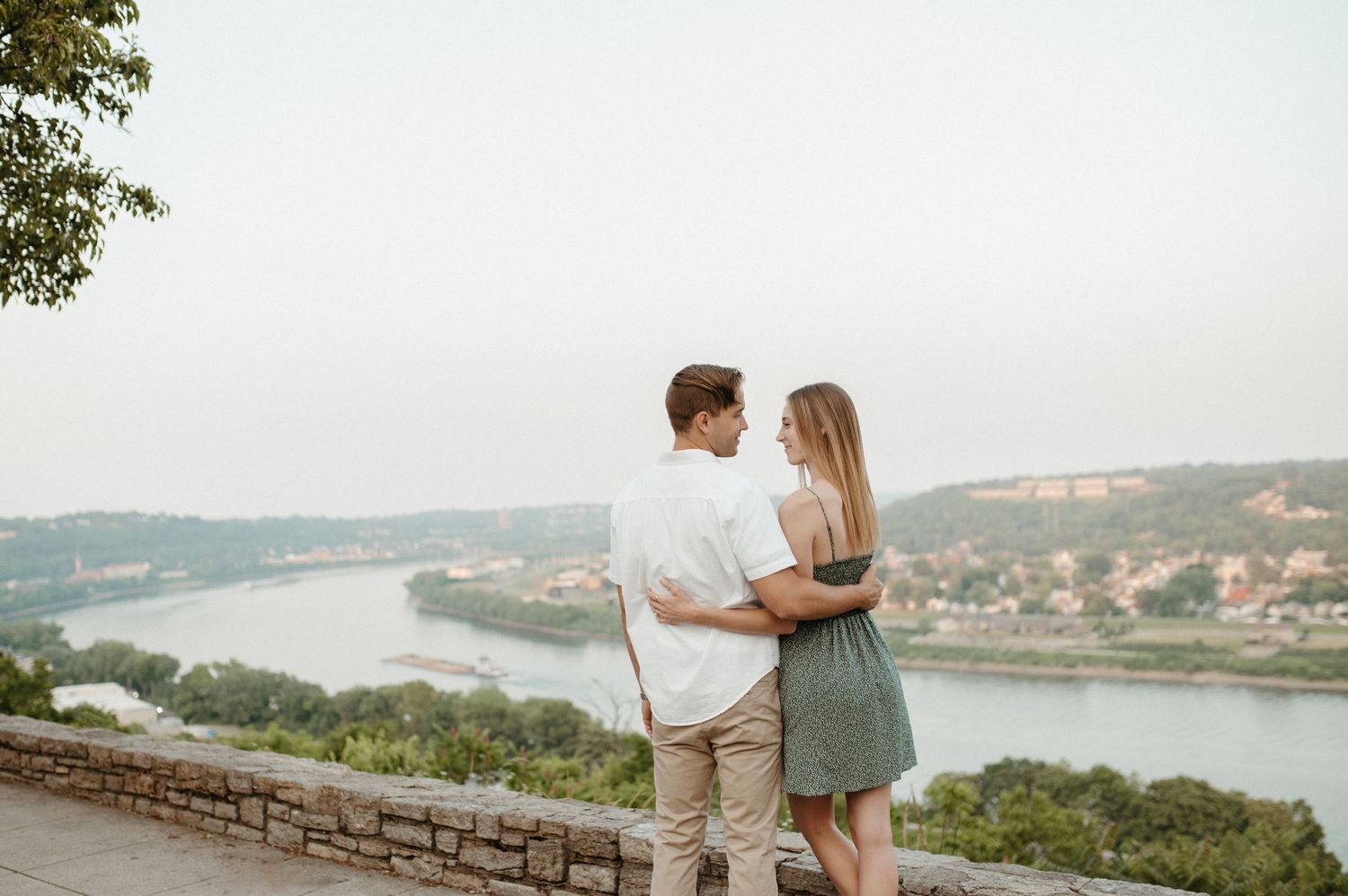 Couple stands together against stone wall overlooking scenic river landscape at sunset.
