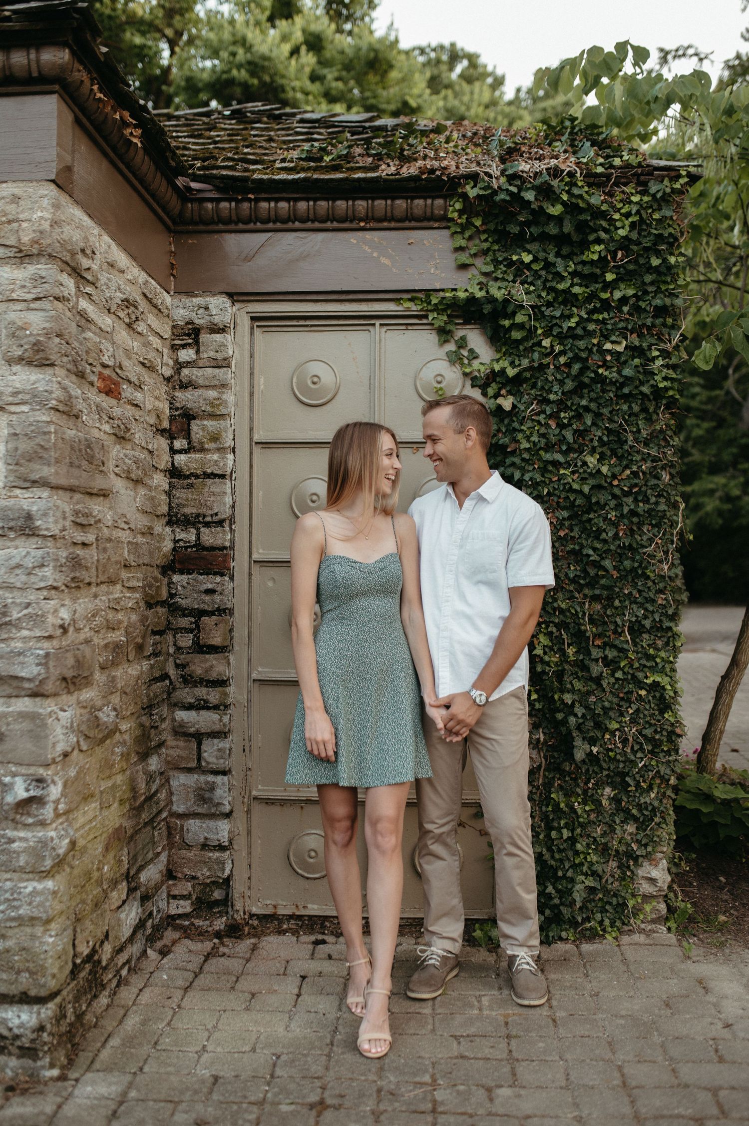 A couple stands in front of an ivy-covered stone wall with ornate door, sharing an intimate moment.