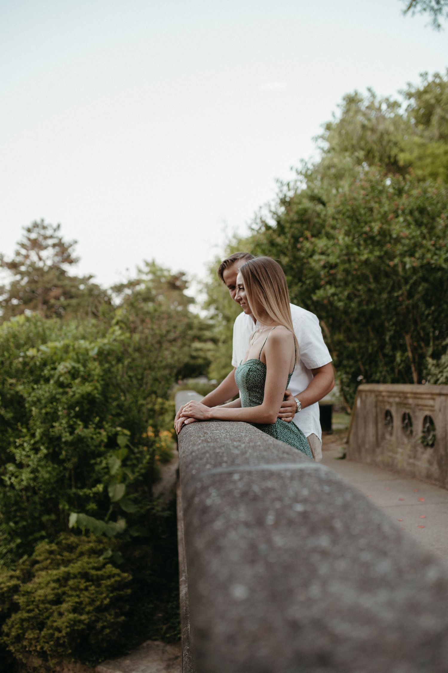 Couple leaning against stone balcony railing overlooking scenic wooded area.