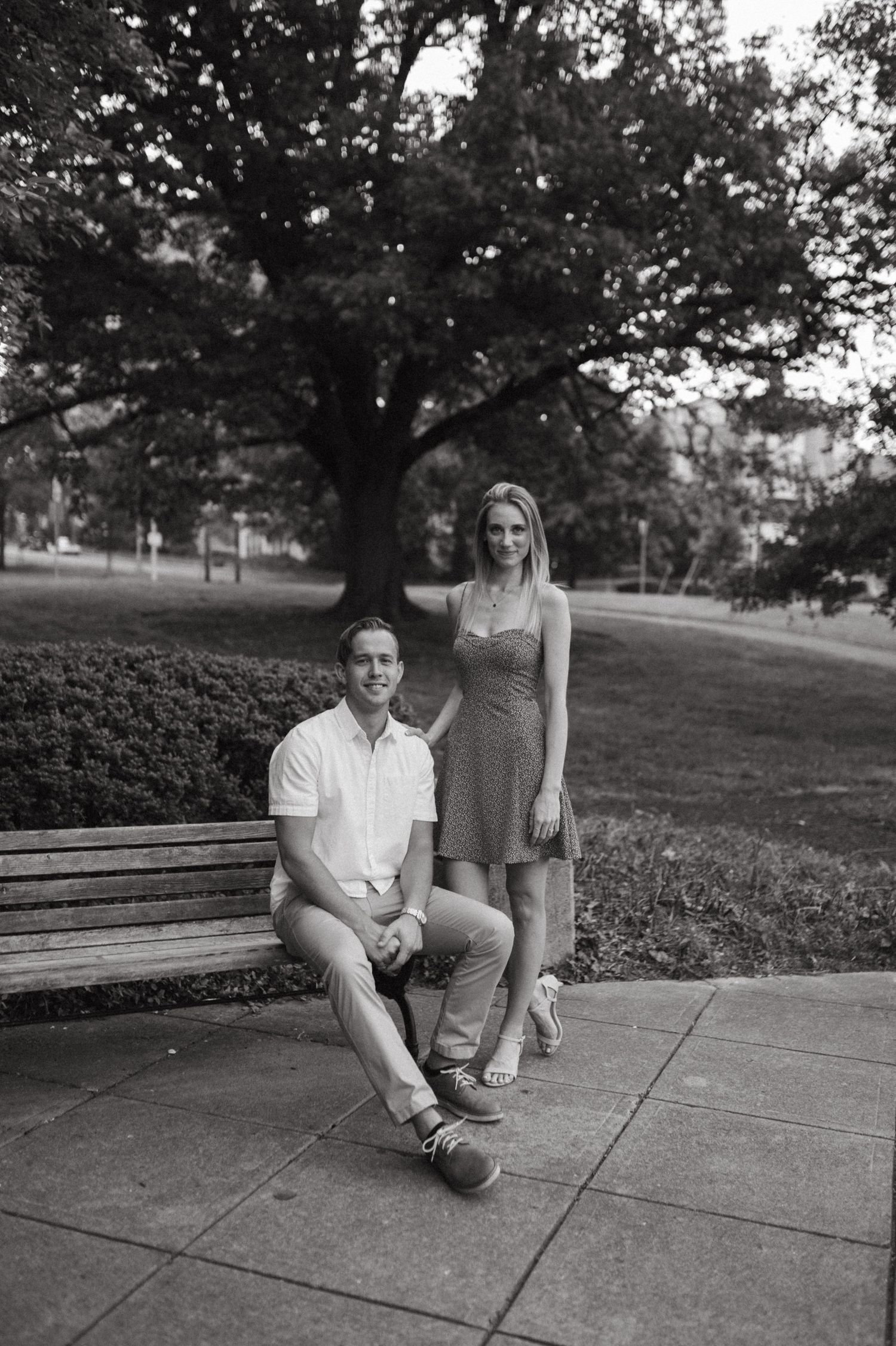 Black and white portrait of couple posing casually by park bench under large tree.