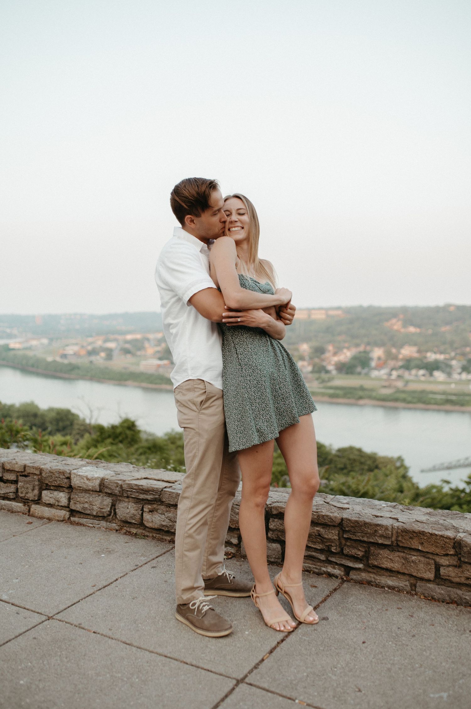Intimate moment between couple standing on stone wall with scenic river view in background.