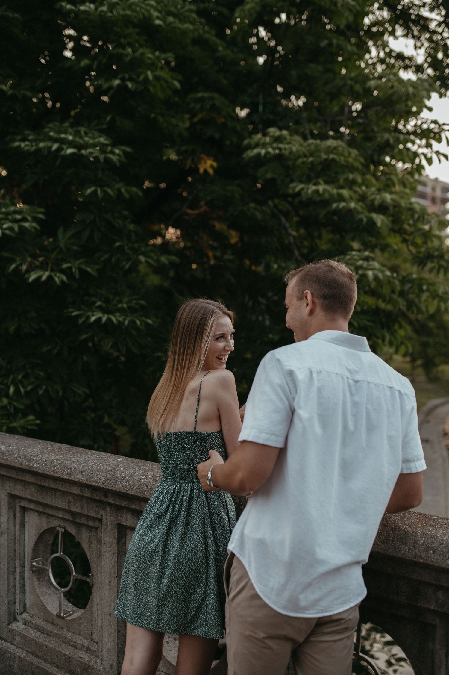 Couple sharing conversation on stone bridge balcony surrounded by green trees.