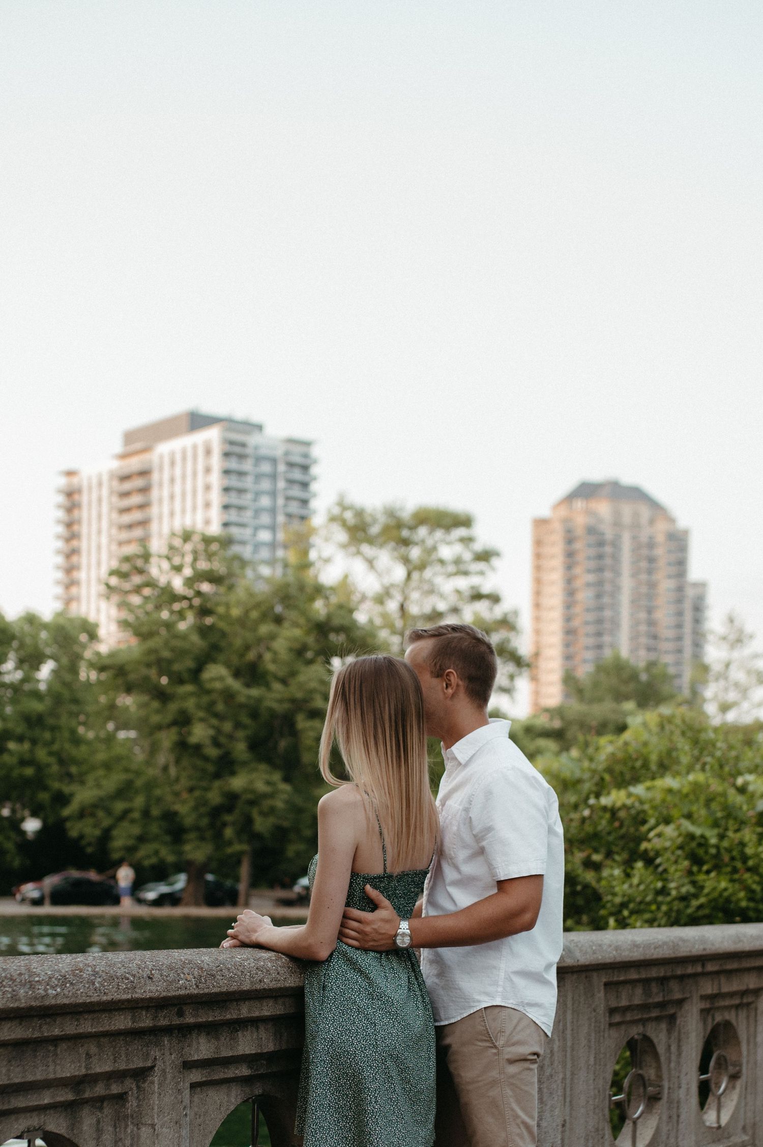 Couple sharing romantic moment on stone overlook with city buildings visible in background.