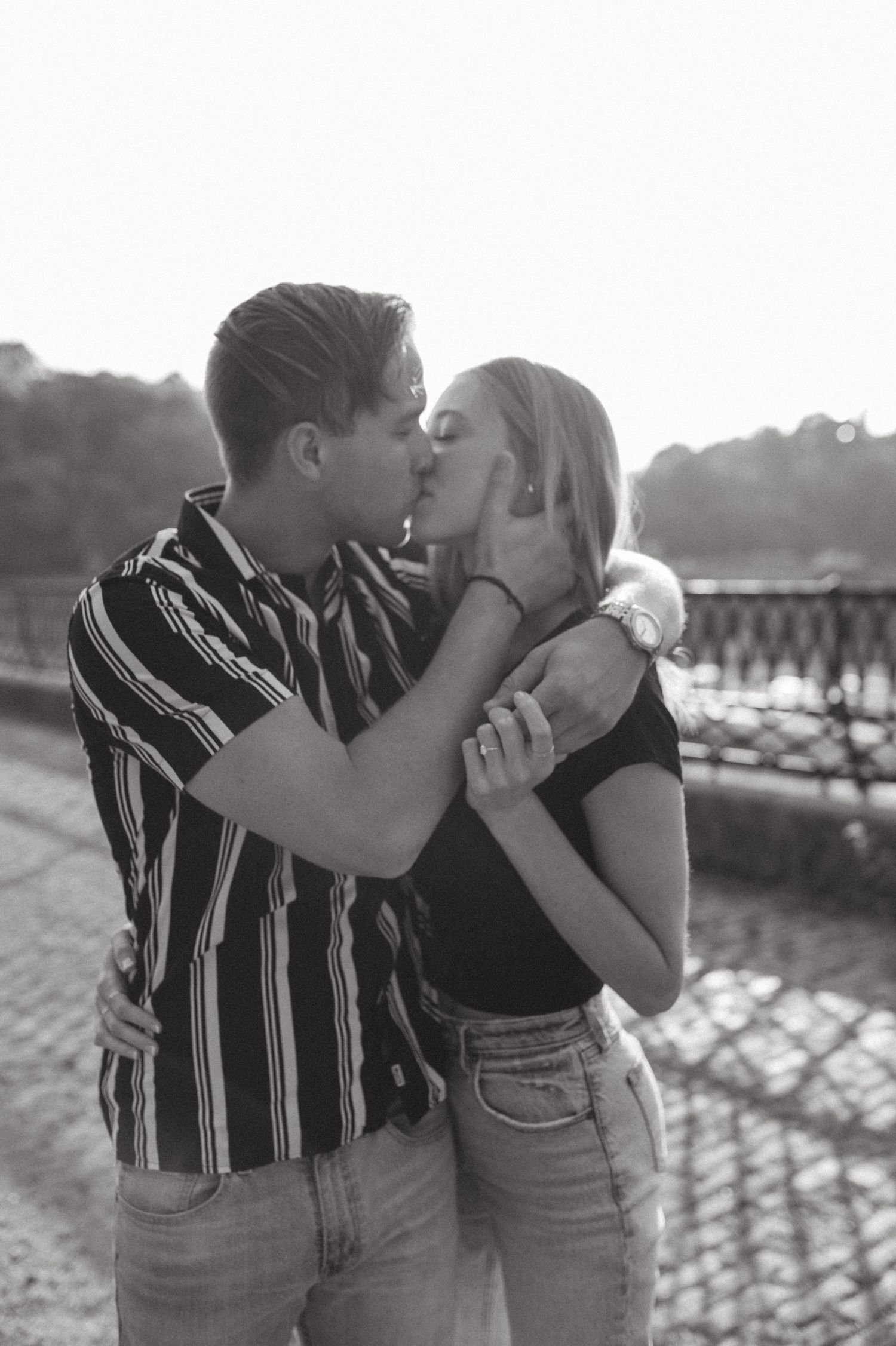 A romantic black and white photo of a couple kissing outdoors against a scenic backdrop with a railing.