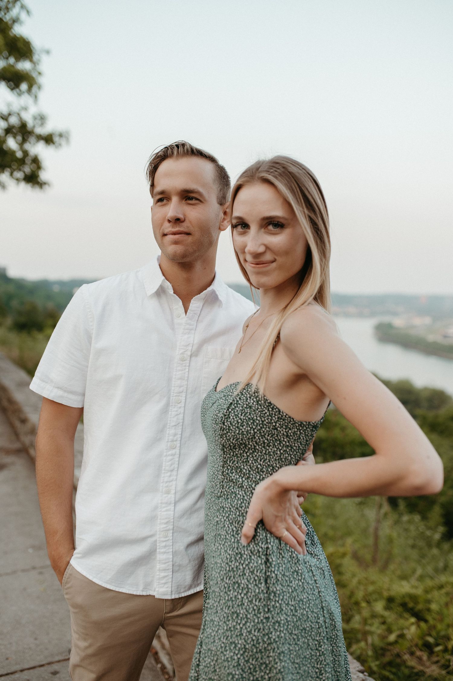 A couple poses together on an overlook during sunset wearing summer outfits with a river view in the background.