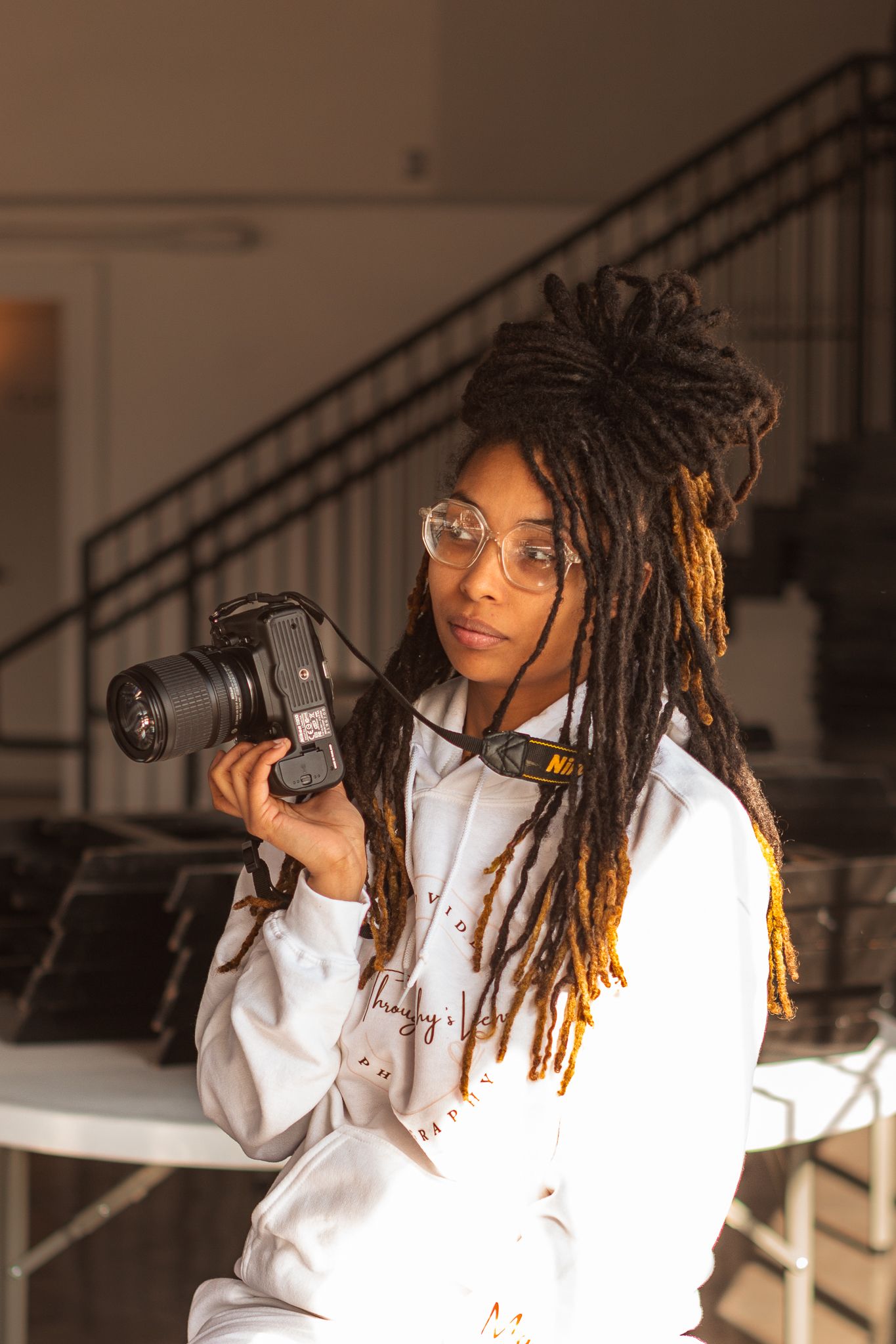 A photographer with locs wearing glasses and a white hoodie holds a DSLR camera.