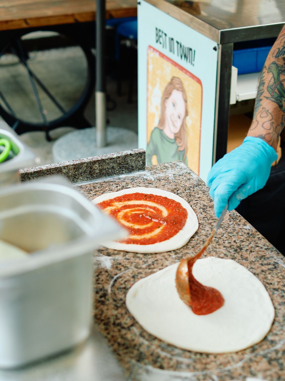 Spreading tomato sauce in circular motion on pizza dough with blue gloved hands.