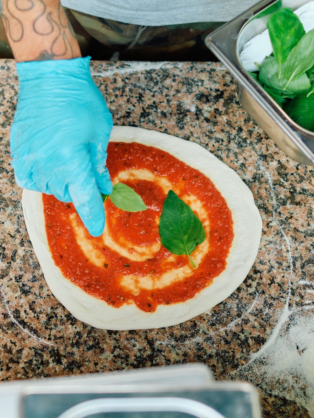 Chef in blue gloves adding basil leaves to pizza sauce spread on raw pizza dough on a granite countertop.