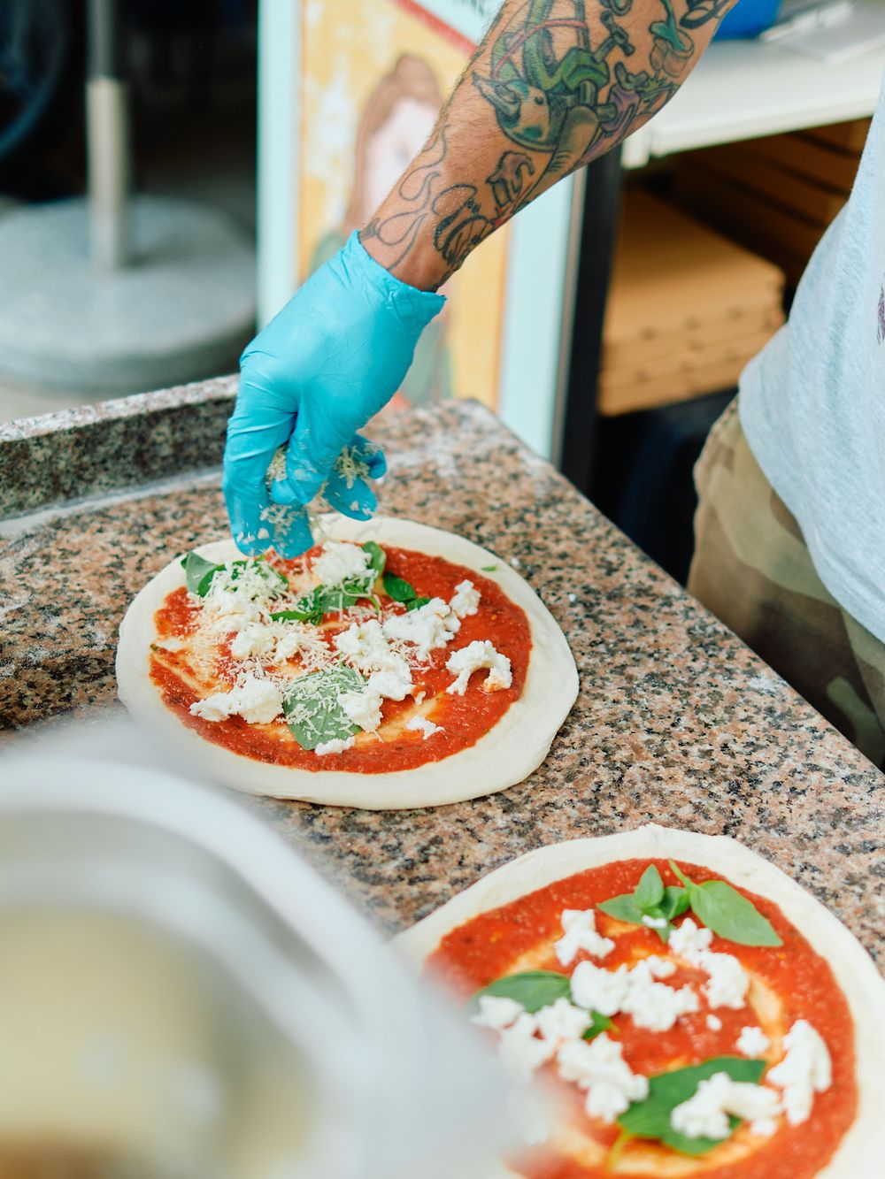 Blue gloved hands sprinkling cheese over pizza sauce and fresh basil leaves.