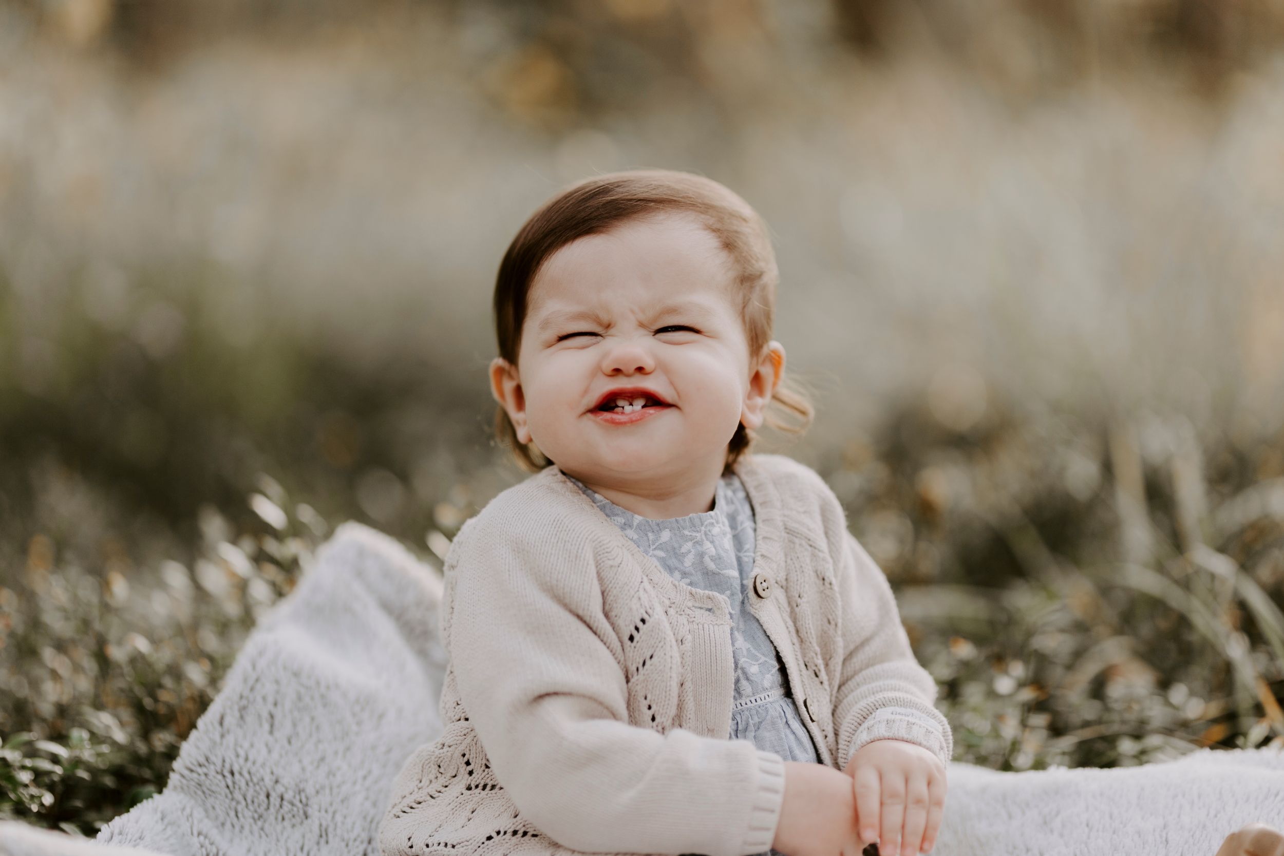 A baby in a beige sweater smiling while sitting outdoors in soft lighting.