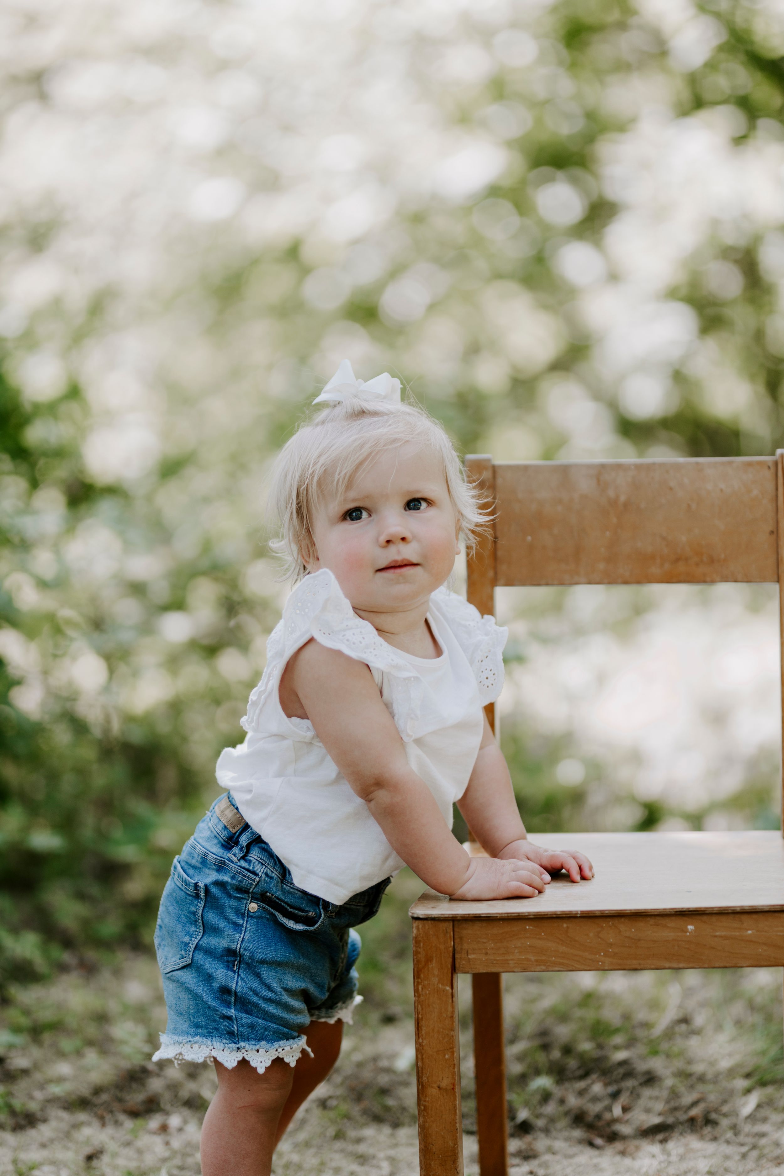 A small child in denim shorts leans against a wooden chair outdoors with bokeh lighting effects.