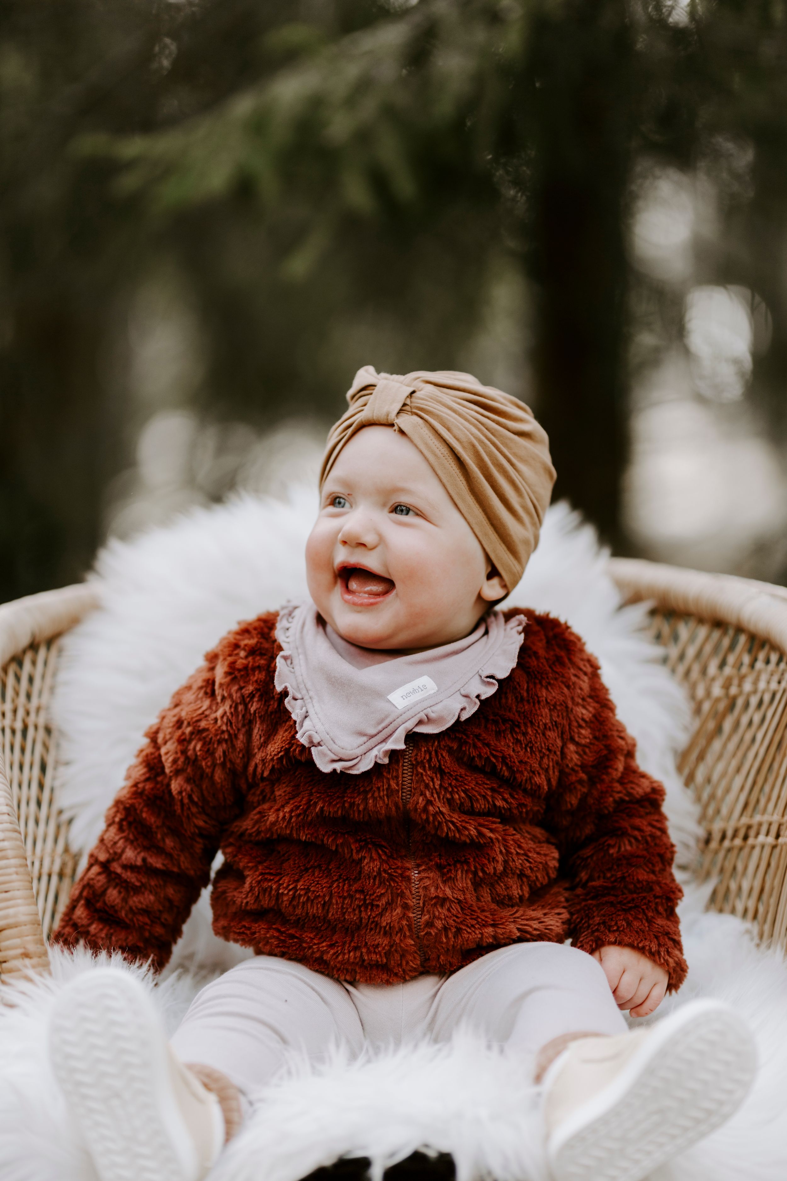 A baby wearing a rust-colored sweater and beige turban sits smiling in a wicker chair.
