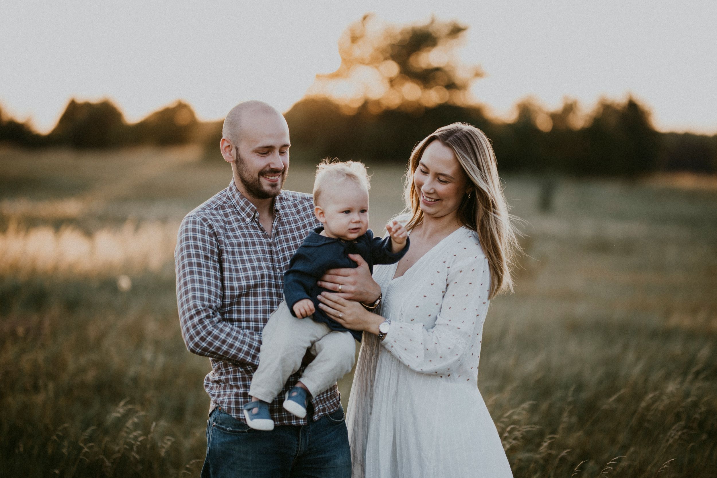 A family of three poses together in a golden sunset field wearing coordinated neutral colors.