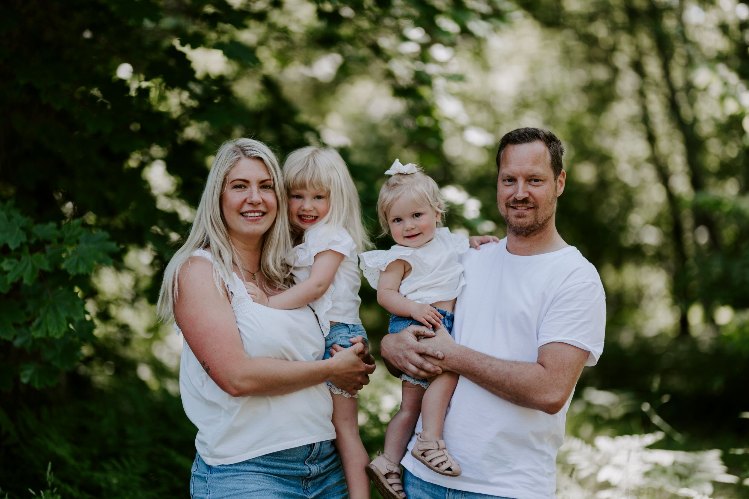 A family of four poses together outdoors against a natural wooded backdrop in casual summer attire.