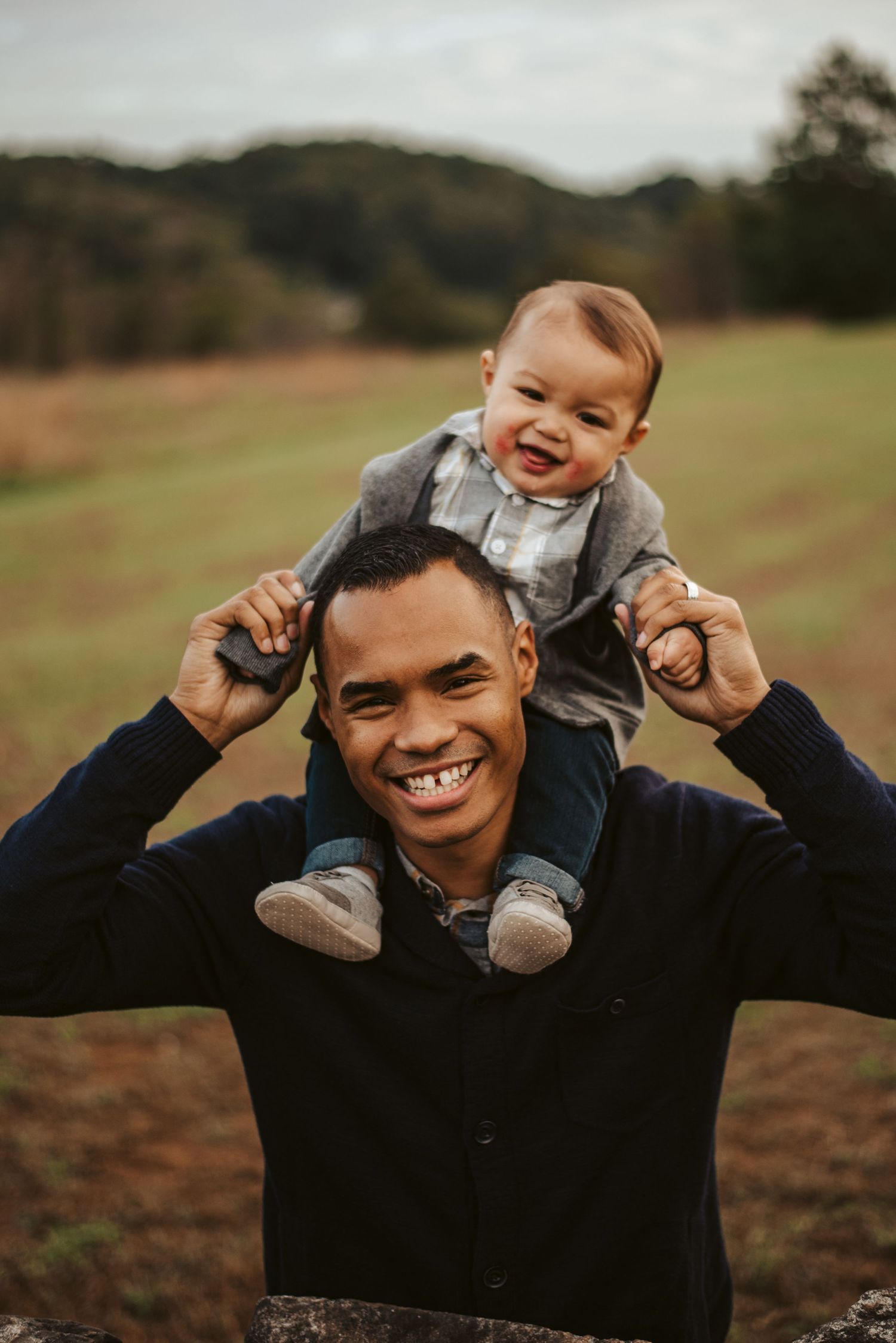 Baby son sitting on father's shoulders, both laughing at the camera, standing in the middle of a field.