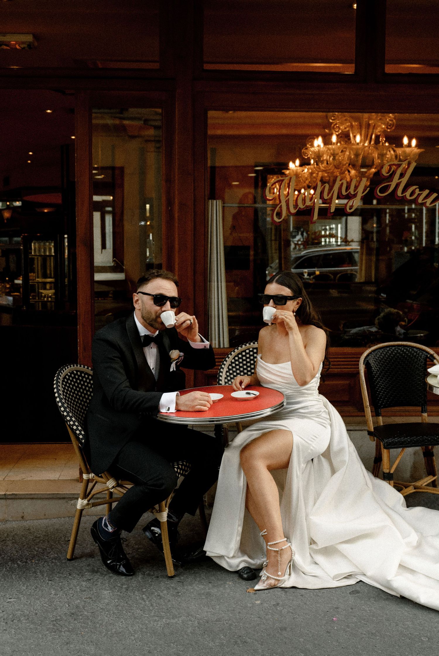 A stylish couple enjoys drinks at a classic Parisian cafe in wedding attire.