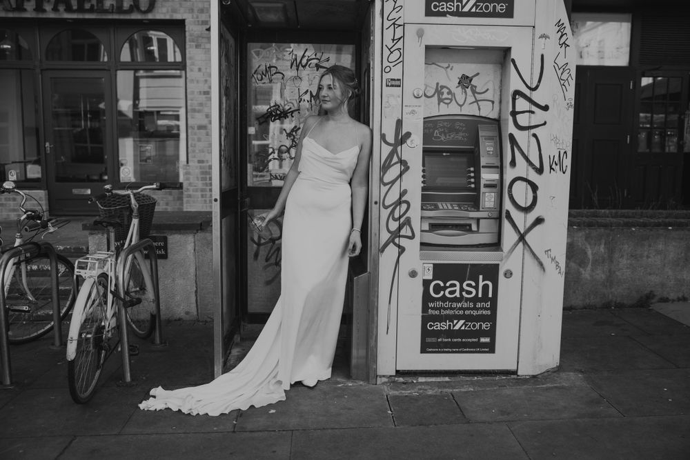 A black and white photo of a model leaning on a phone box drinking a coffee
