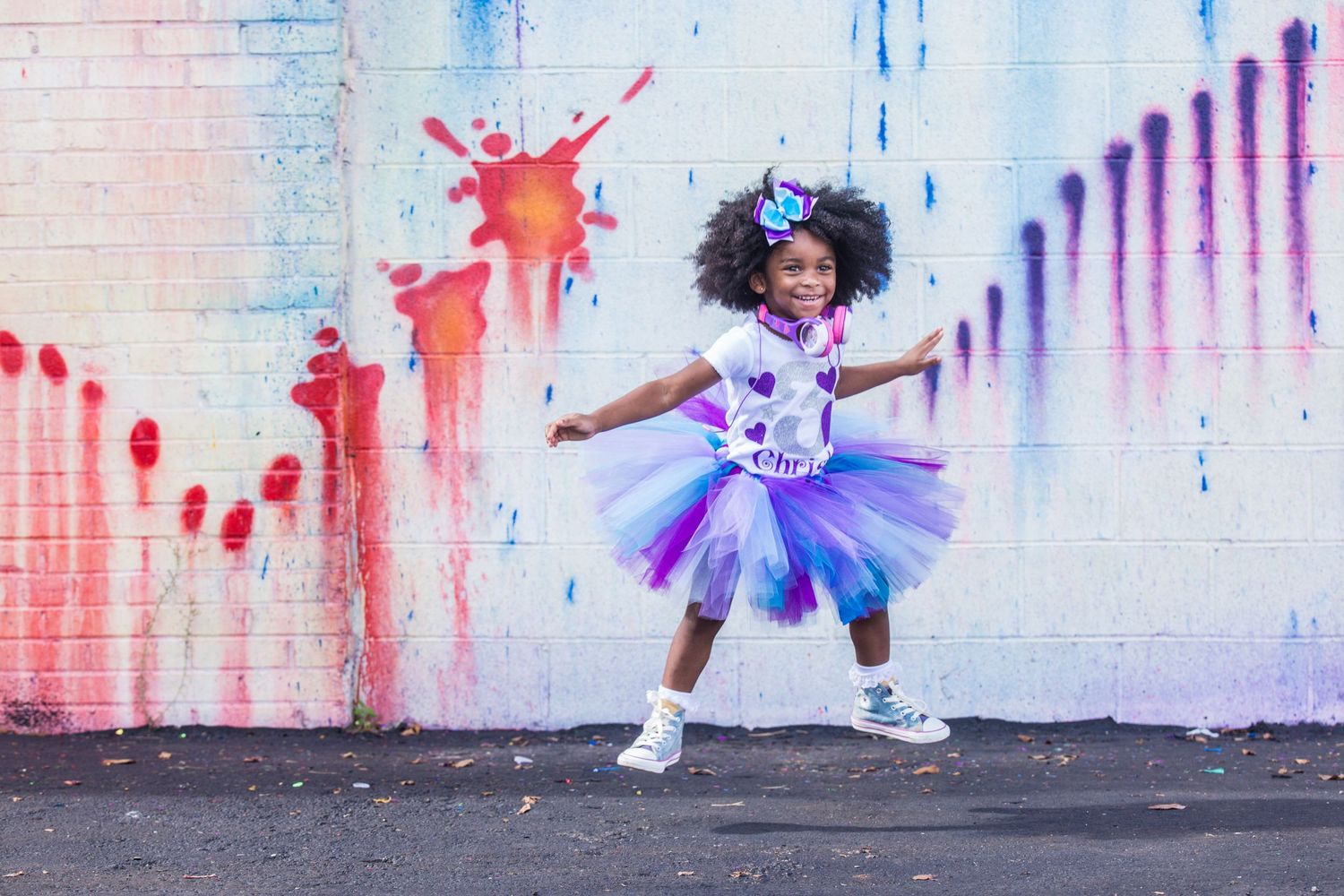 A young dancer in a purple tutu and white sneakers poses joyfully against a colorful paint-splattered wall.