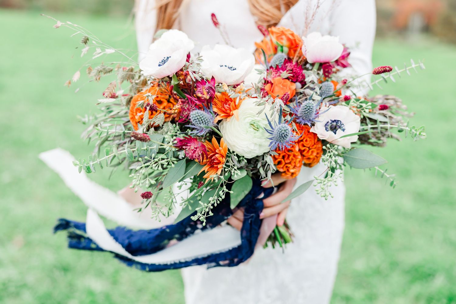 A vibrant autumn-themed wedding bouquet with orange and white flowers held against a grassy outdoor background.