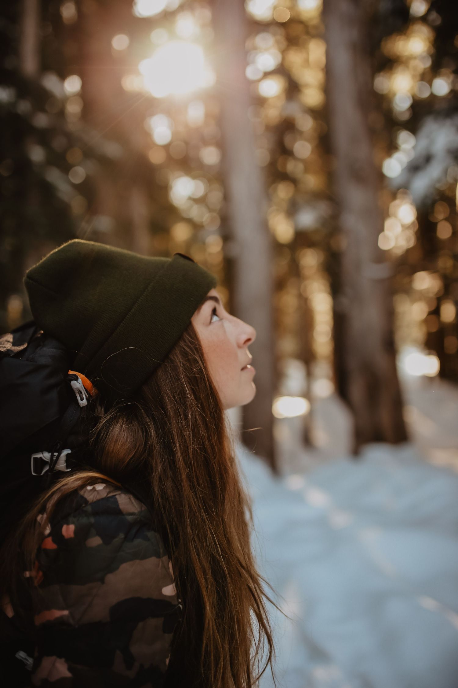 A winter hiker gazes upward in a snow-covered forest during golden hour.