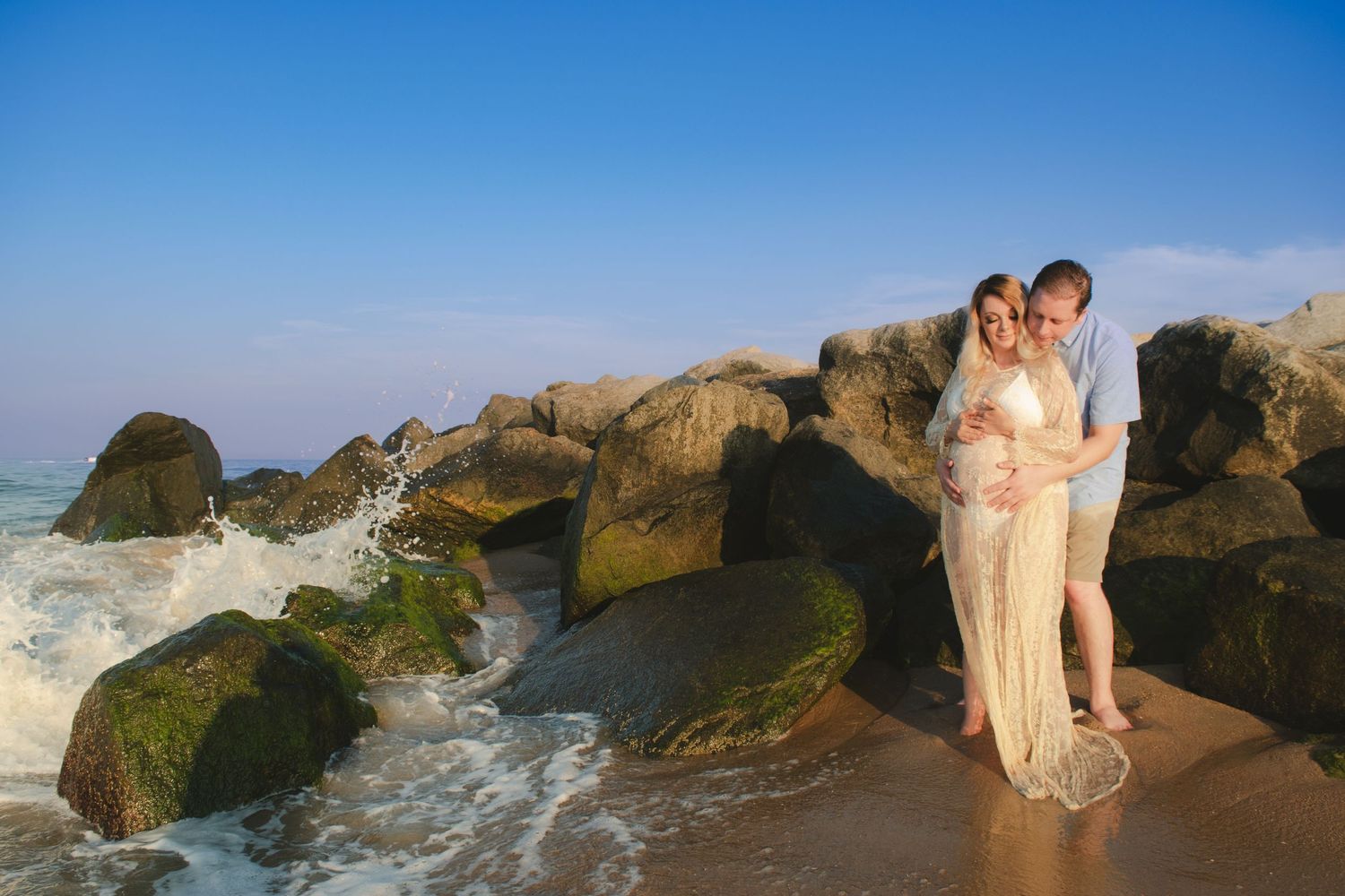 Couple standing on rocky beach at sunset with waves crashing.