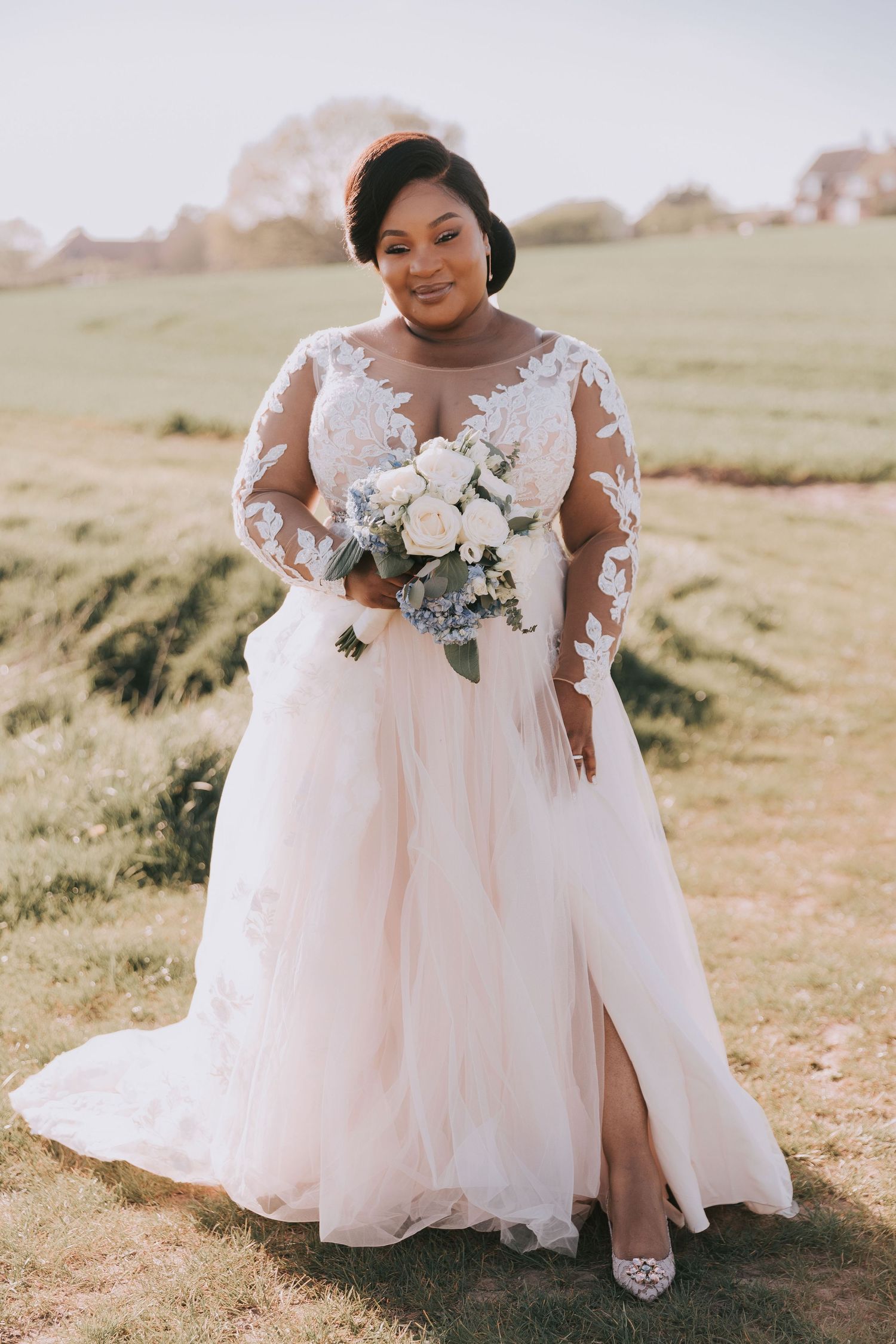 Bride in lace gown poses with bouquet in sunlit outdoor setting.