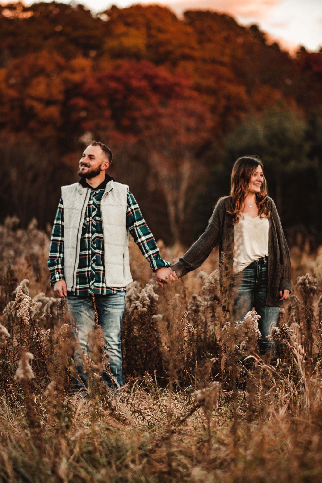 A couple holds hands while walking through a golden autumn field at sunset.