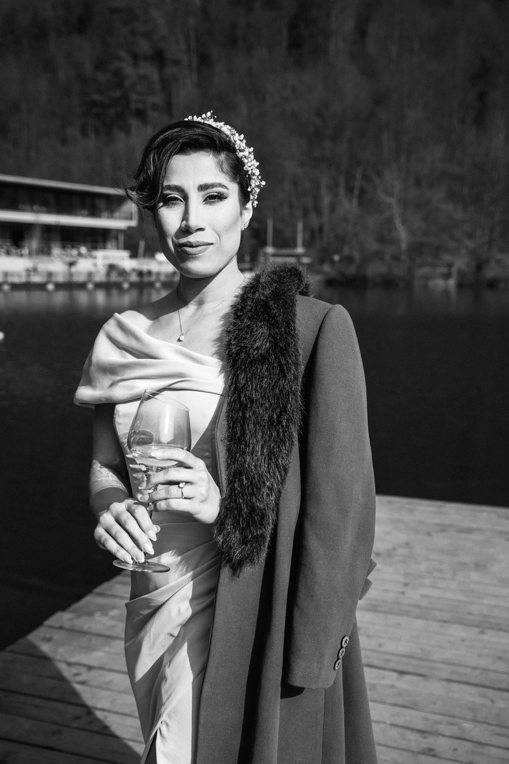 Elegant bride in white satin dress and fur stole poses on wooden deck by a lake.