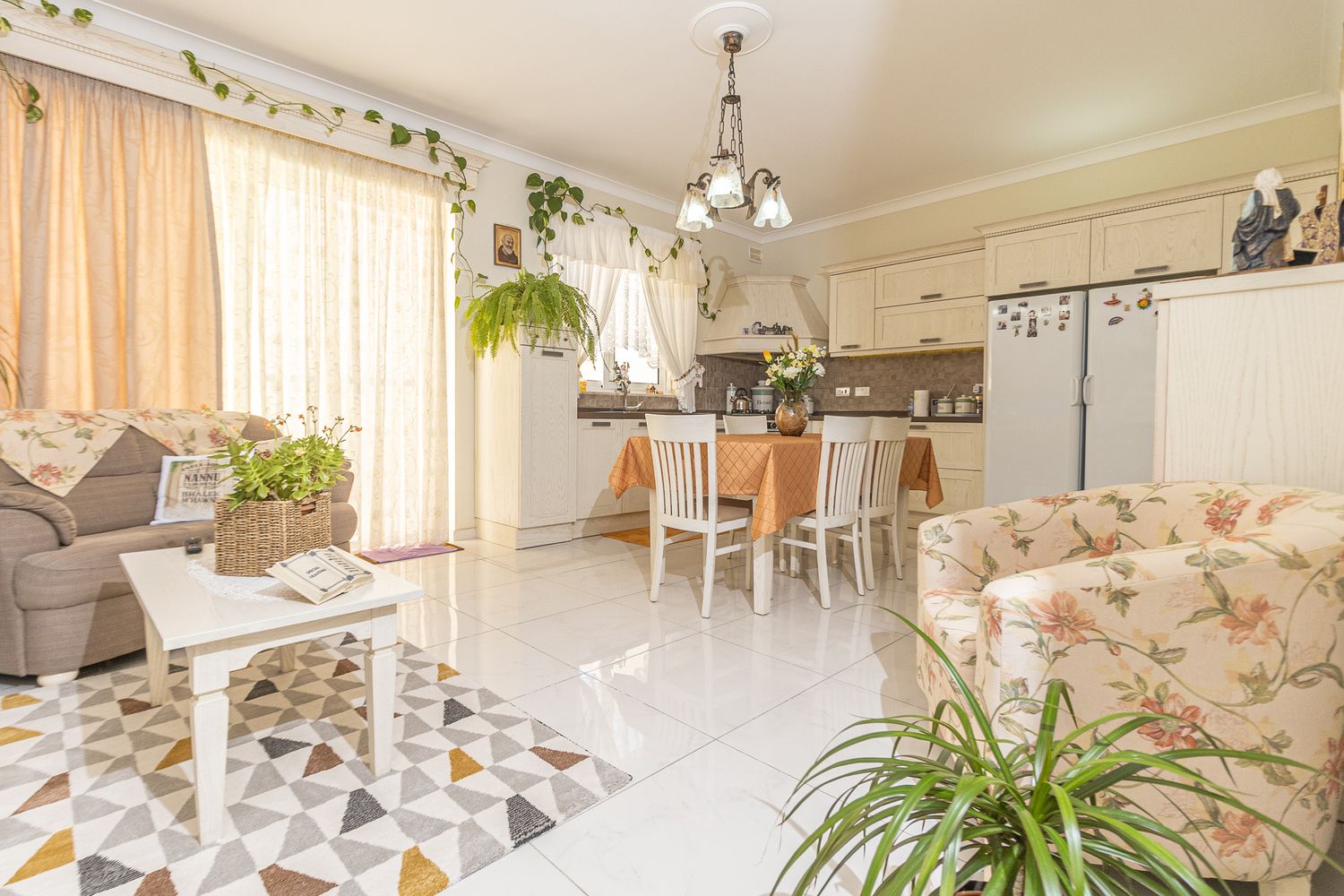 Bright cozy living room with floral sofas, dining area, plants, and white tiled floors.