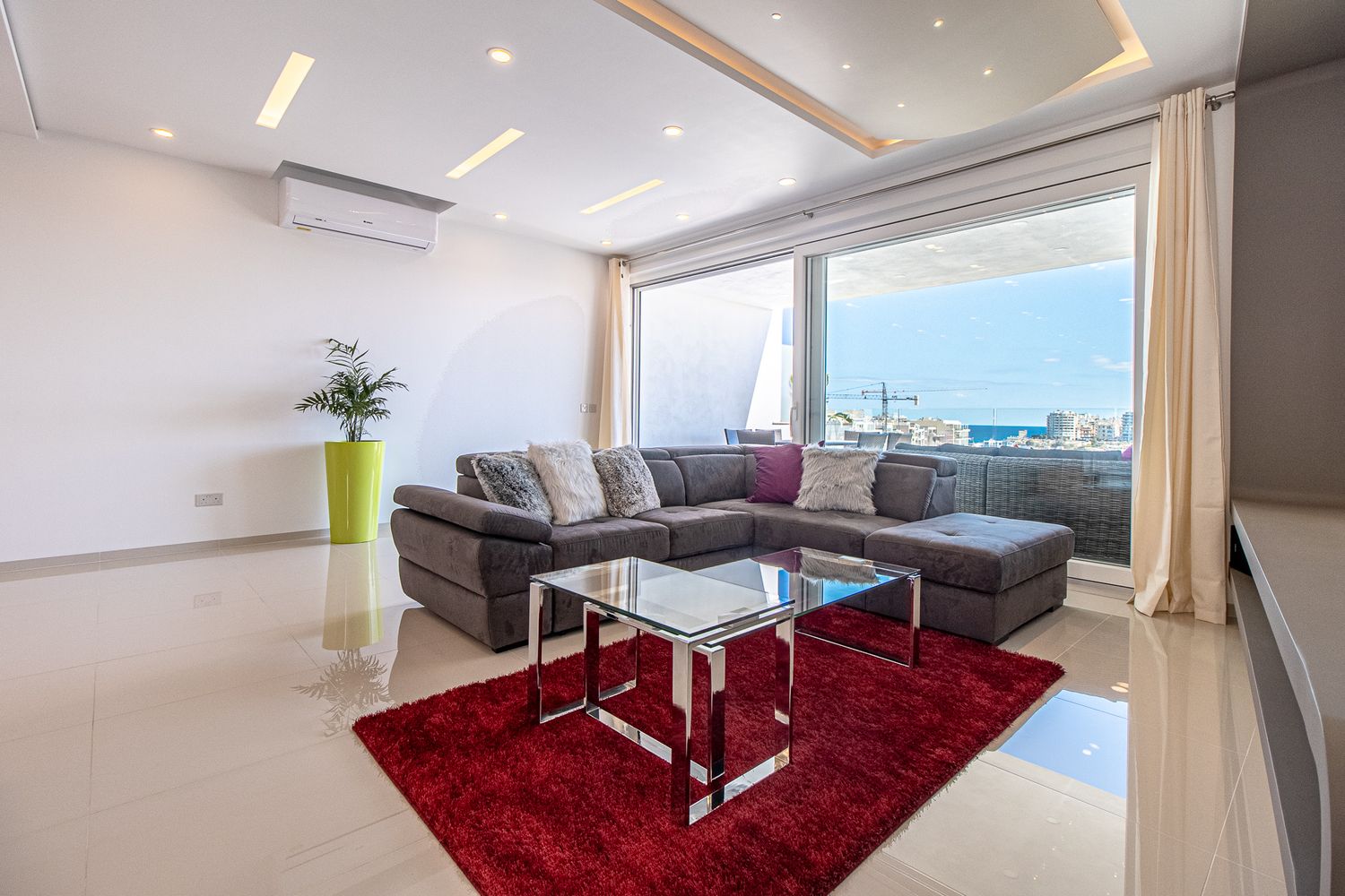 Modern living room with gray sectional sofa, red rug, glass coffee table, and panoramic city view windows.