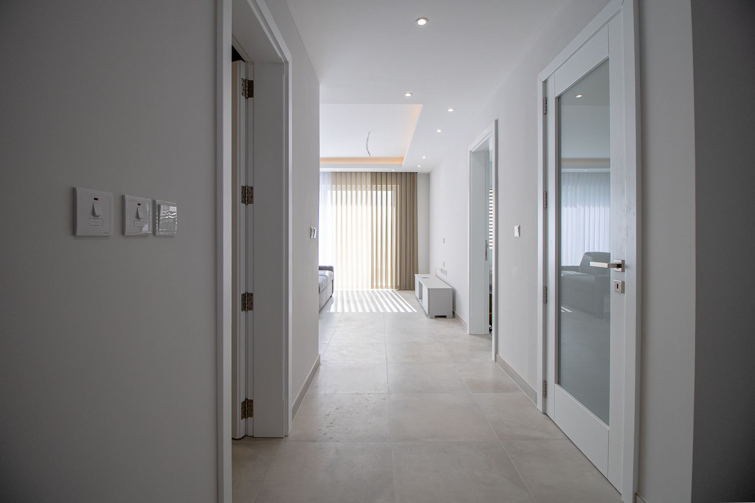 Bright minimalist hallway with white doors, recessed lighting, and neutral tile flooring in a modern home.