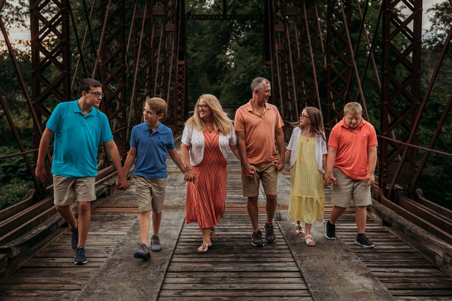 Family walking together on a wooden bridge surrounded by tall trees.