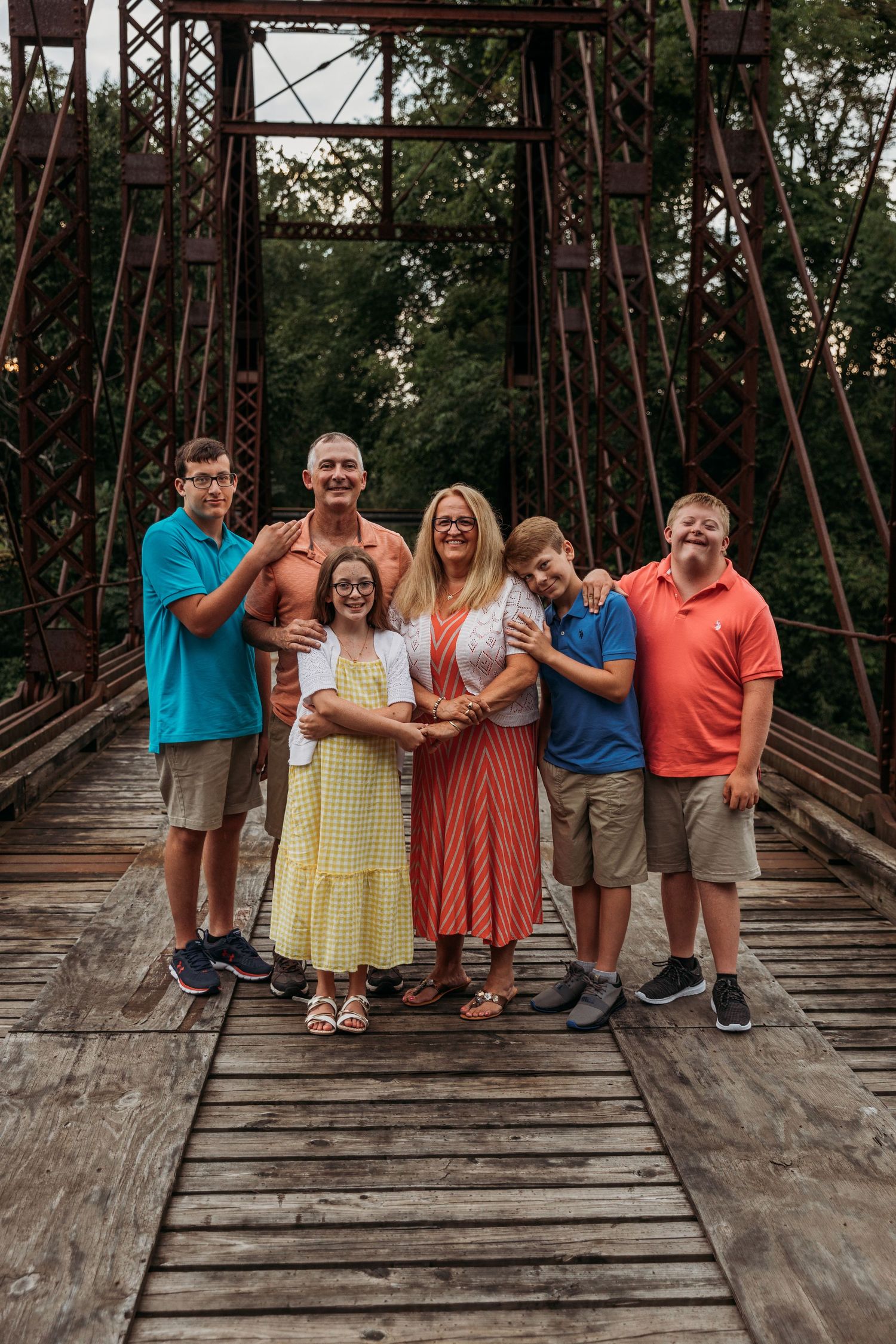 Family wearing colorful casual clothes poses together on rustic wooden bridge.