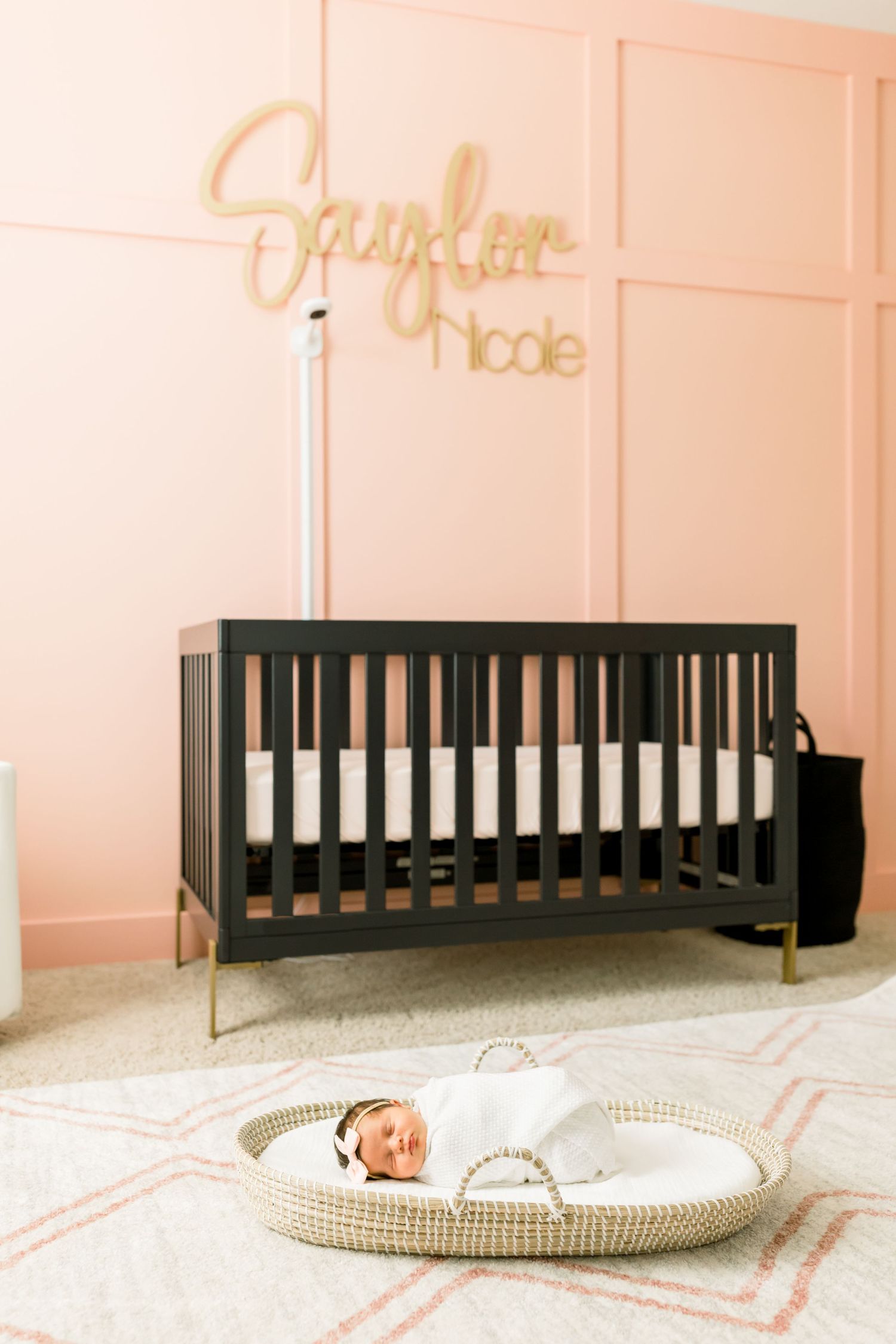 Modern nursery with black crib, gold name sign, and woven basket on pink patterned carpet.