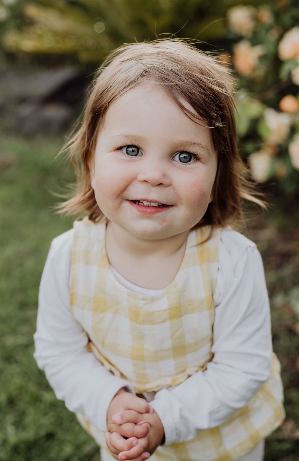 Portrait of a smiling toddler wearing a yellow gingham dress and white long sleeve shirt.