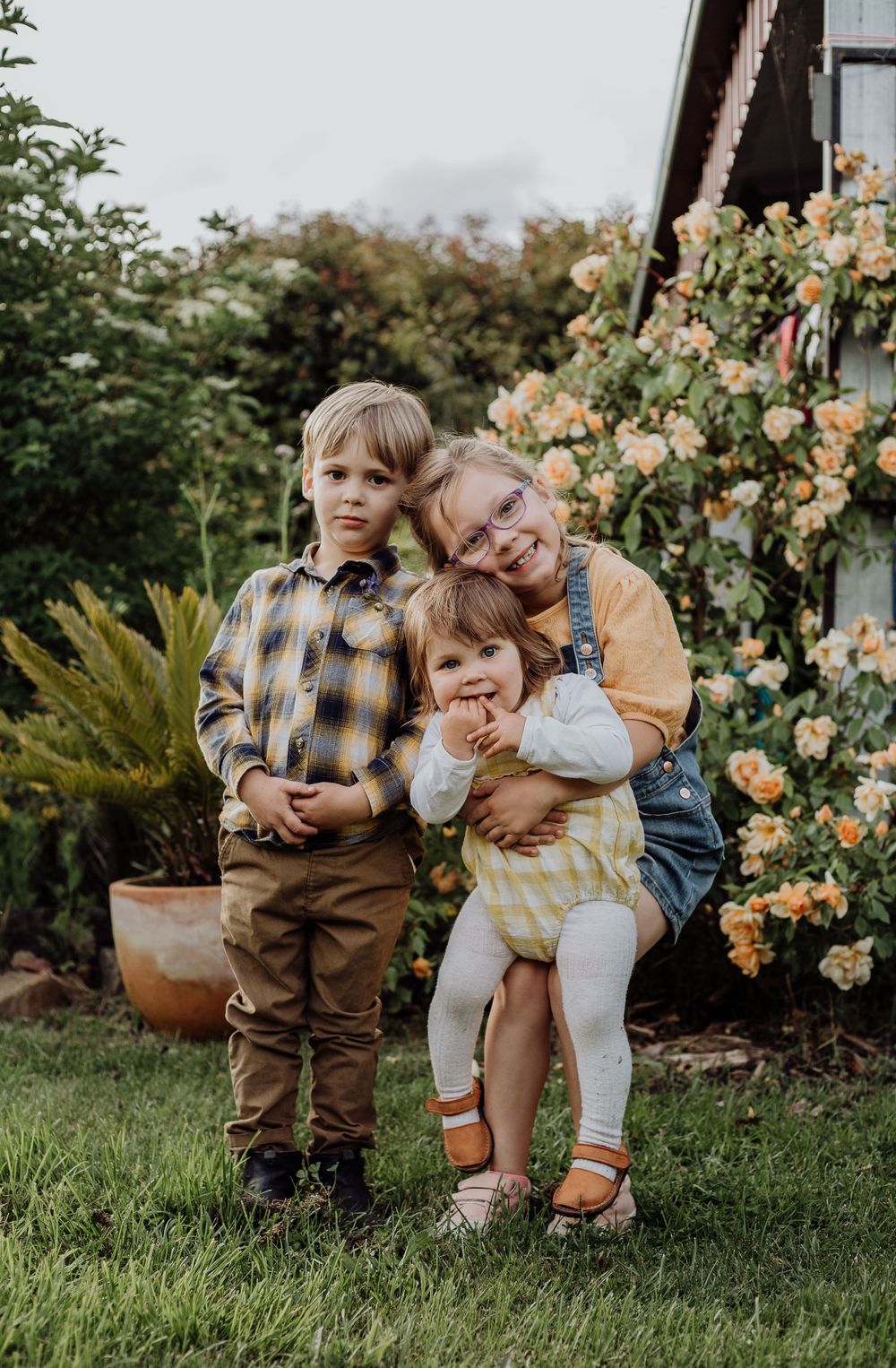 Three young children pose together in front of blooming roses and garden foliage.