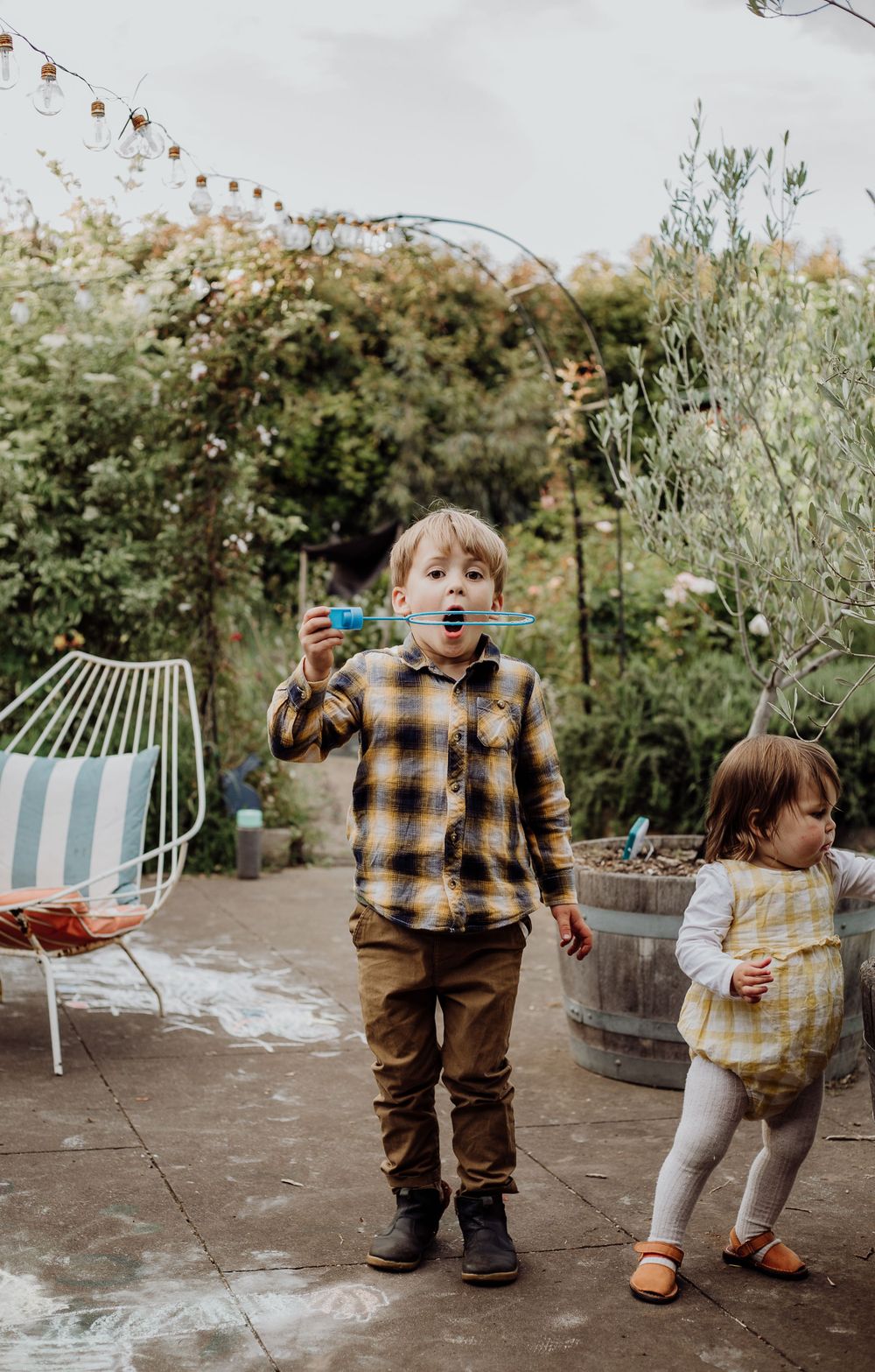 Children playing with bubbles on patio with garden furniture and plants in background.