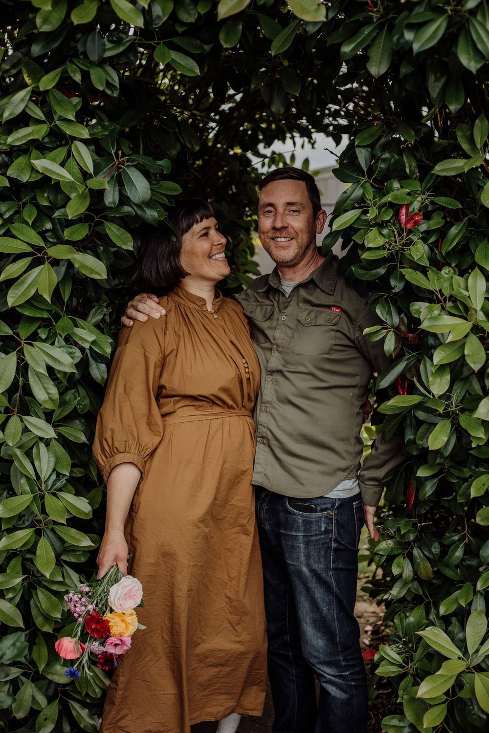 A couple shares a tender moment against a backdrop of green leaves while holding a colorful bouquet.